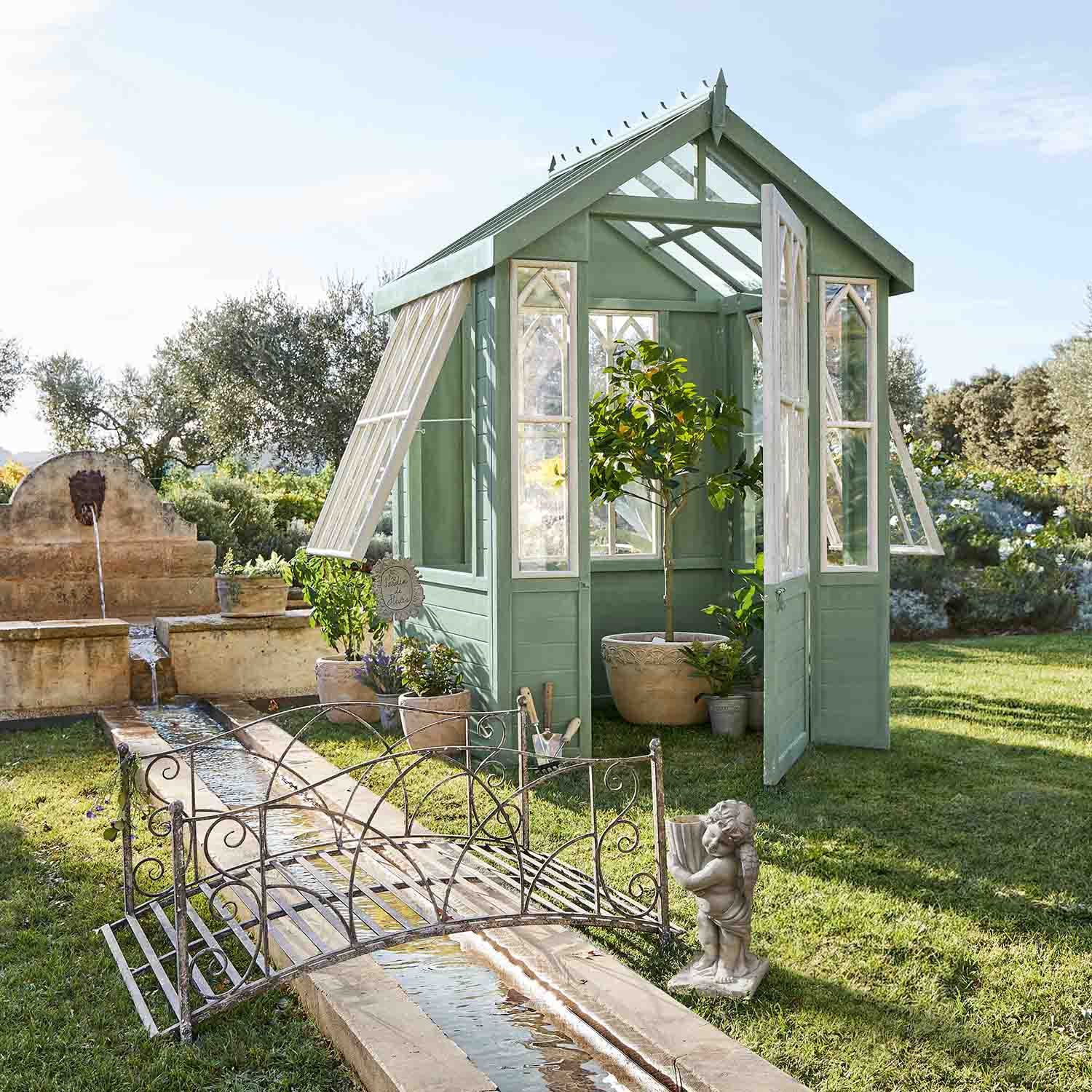 Grüne Holz-Gewächshaus mit weißen Fenstern im Garten mit Pflanzen und Brunnen