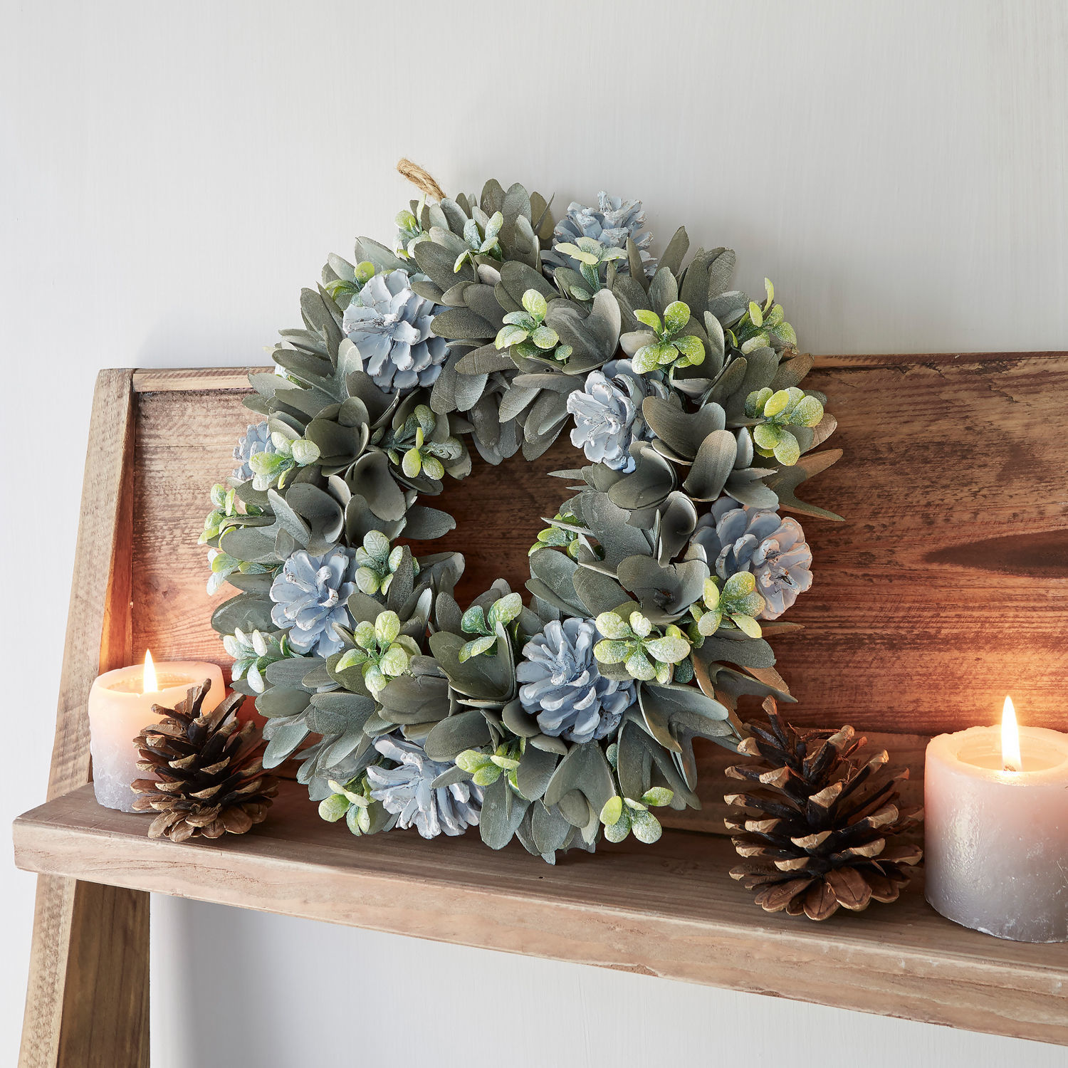 Kranz Deinze Dekorativer Kranz aus grauen Blättern und blauen Zapfen auf Holzregal mit Kerzen und Kiefernzapfen