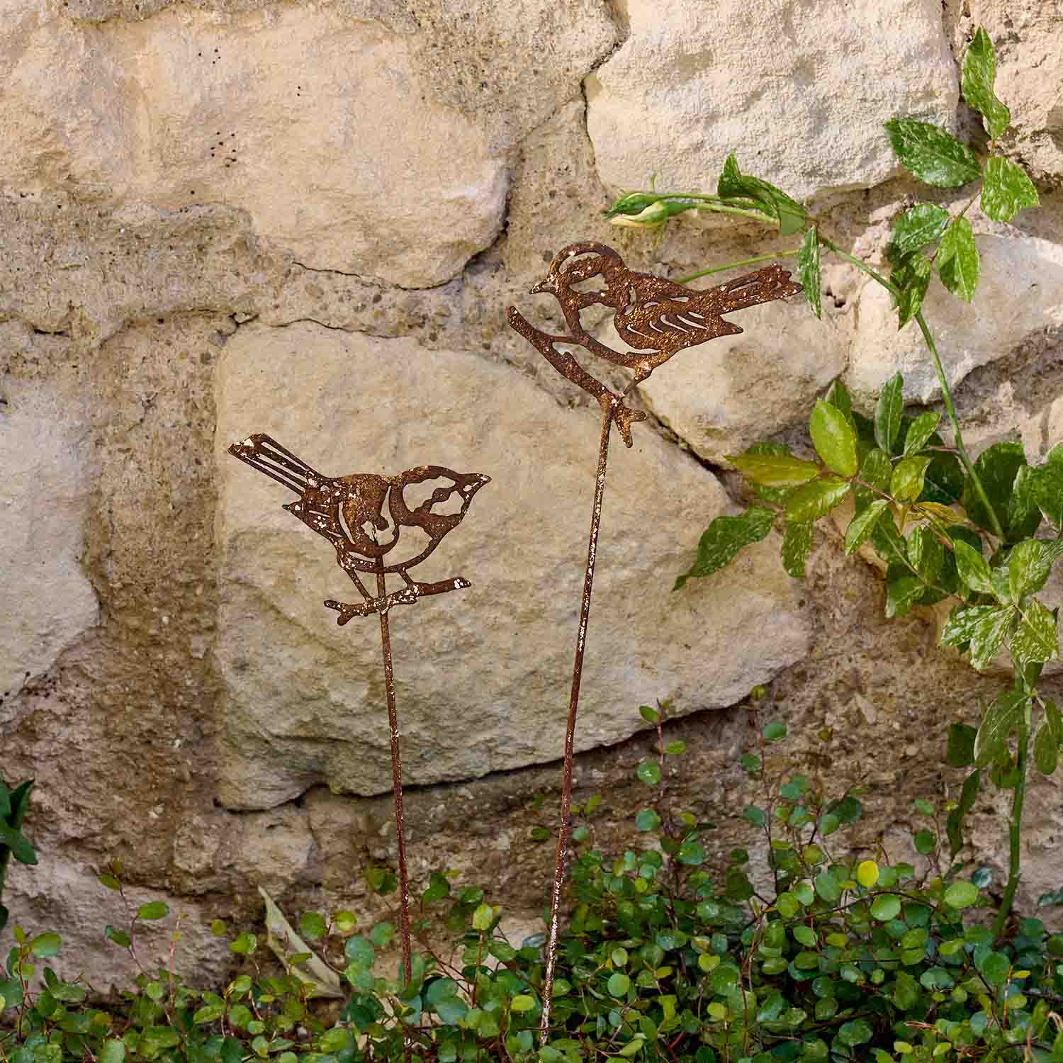 Dekorative Metallvögel in Rostoptik auf Stäben im Garten vor einer Steinmauer