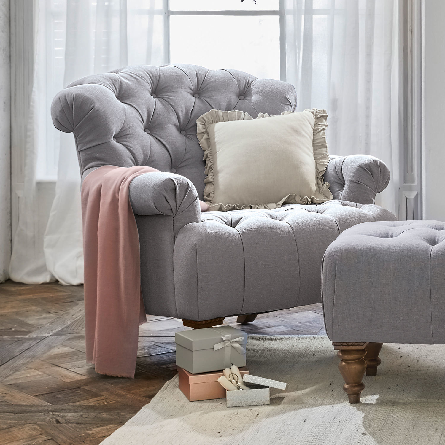 Sessel Springfield Grau gepolsterter Sessel mit Hocker, cremefarbigem Kissen und rosa Decke auf Holzboden im Wohnzimmer