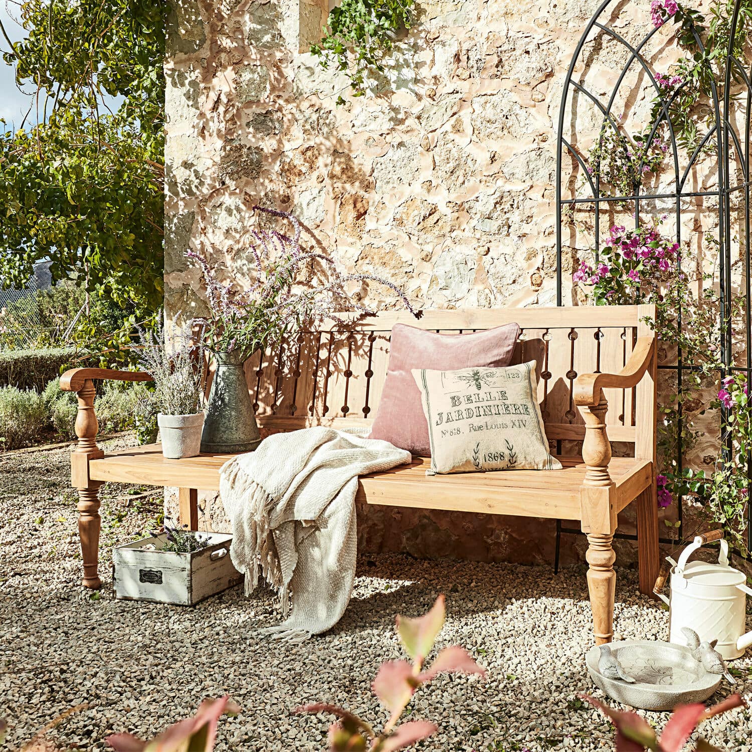 Bank Loudoun Helle Holzbank im Garten mit Kissen und Decke auf Kiesboden vor Steinmauer und Pflanzen
