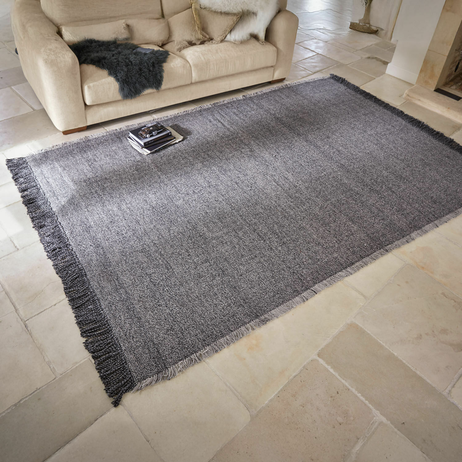 Grauer Teppich mit Fransen auf hellem Boden vor beigem Sofa mit Kissen und Felldeko