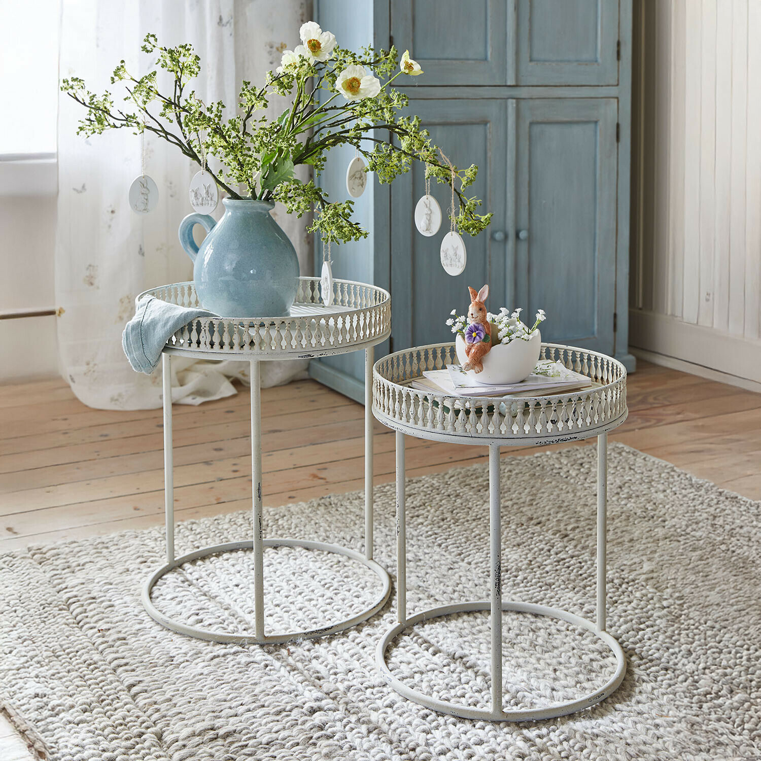 Zwei weiße Metall-Beistelltische mit filigranem Rand auf grauem Teppich, dekoriert mit Vase und Hasenfigur