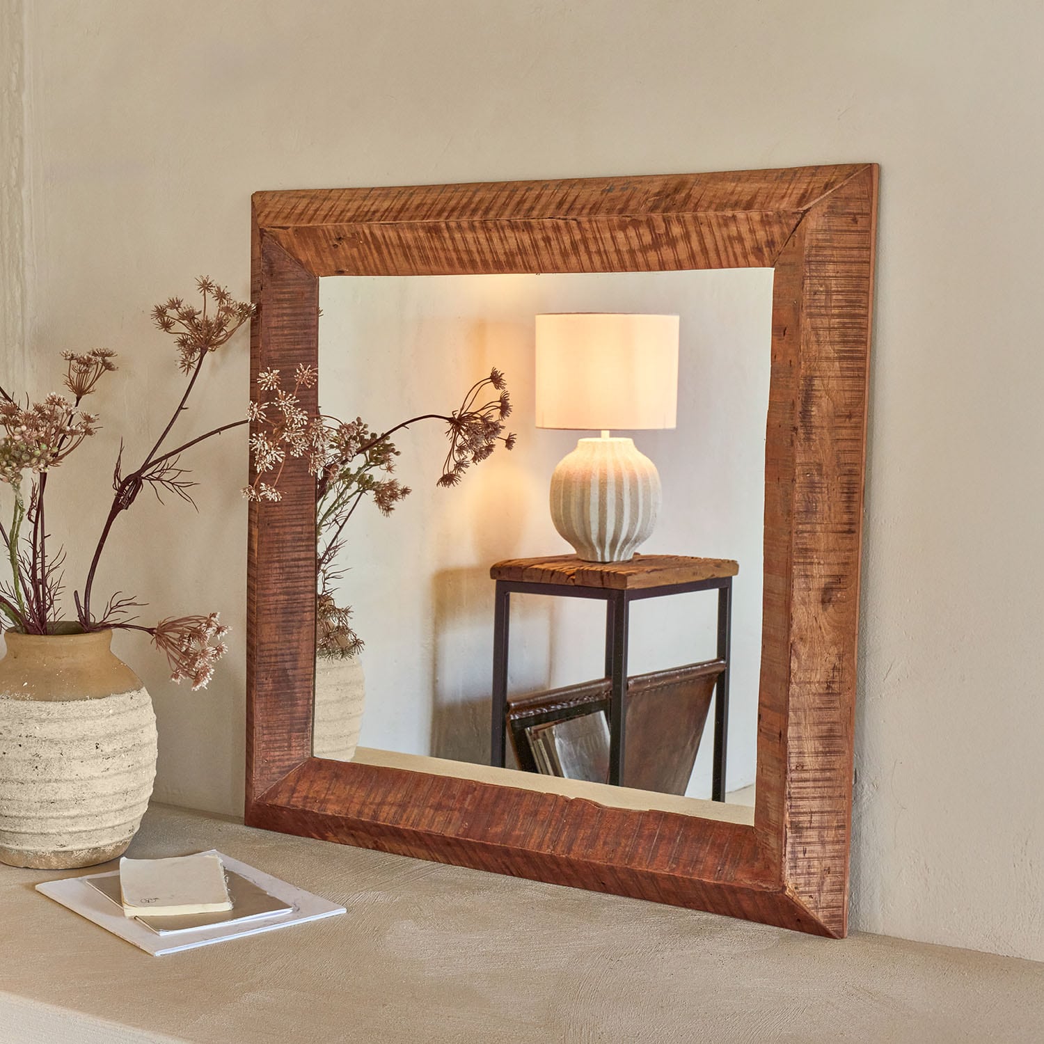 Quadratischer Spiegel mit dunklem Holzrahmen an der Wand, reflektiert Lampe und rustikalen Tisch