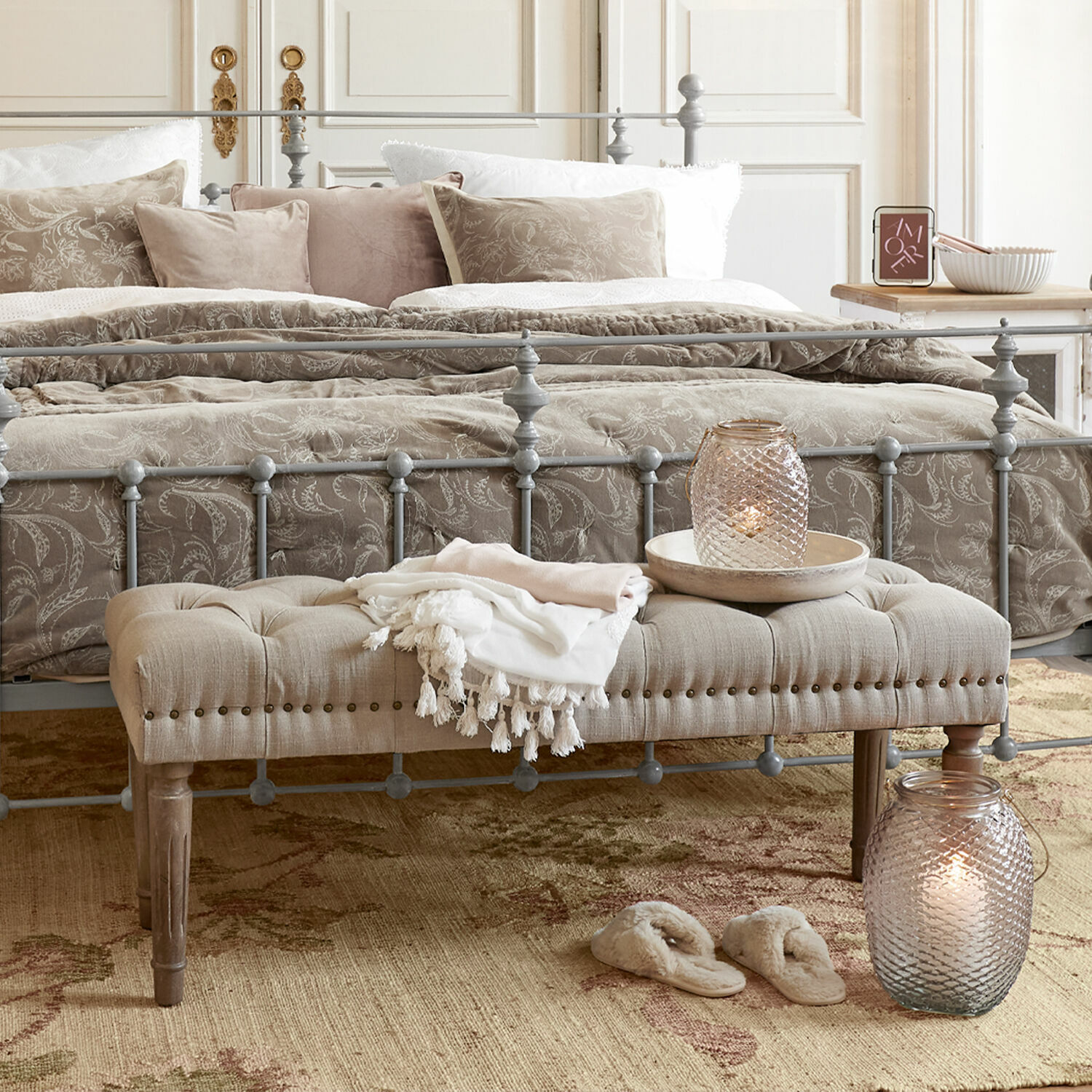 Graues Metallbett mit beige gepolsterter Bank und dekorativen Kissen in gemütlichem Schlafzimmer