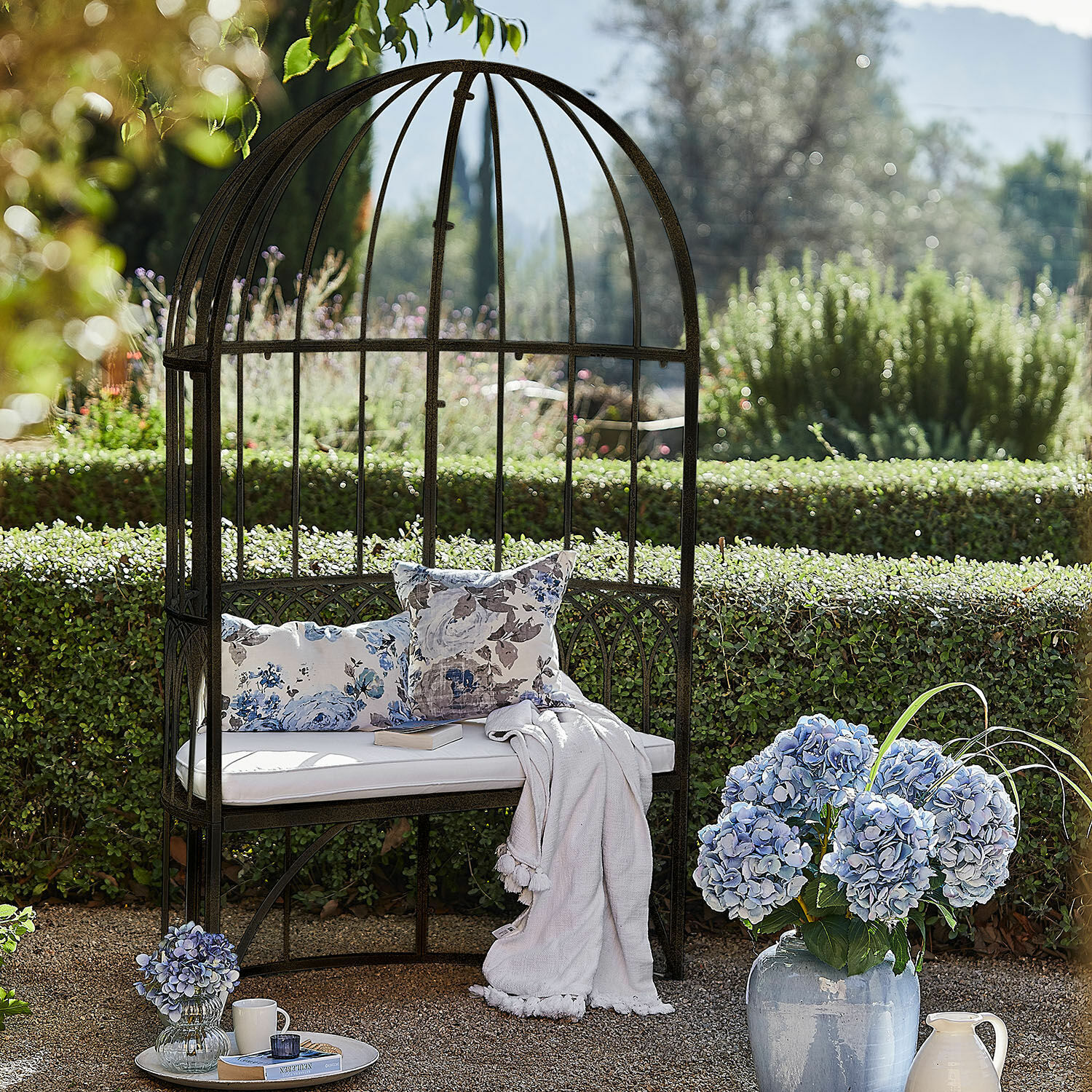 Bank Rivandelle Garten mit Metallbank in Käfigform, weißem Kissen und blauen Blumen in Vase