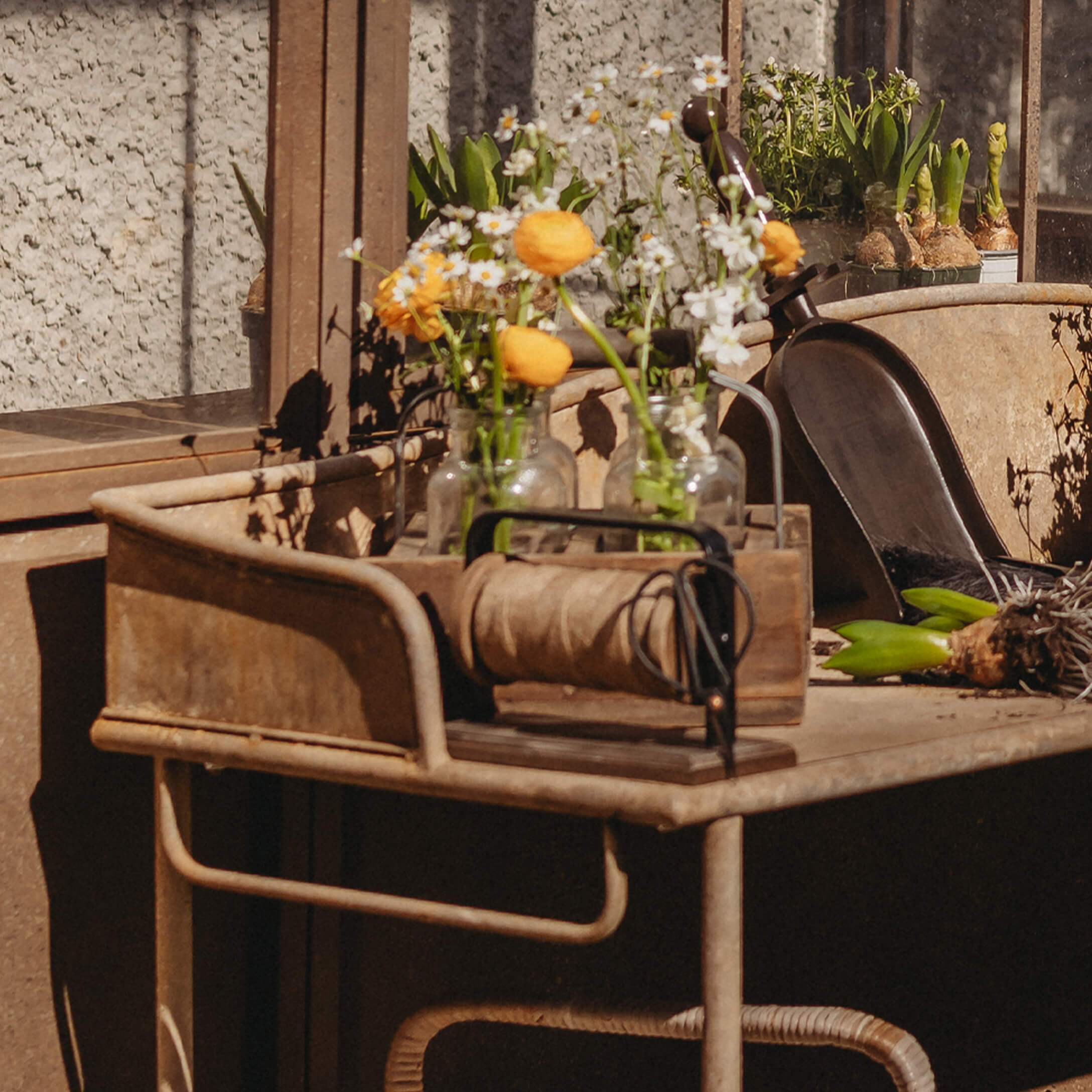 Metalltisch mit Glasvasen und gelben Blumen, Gartengeräte und Seilrolle auf sonnigem Balkon