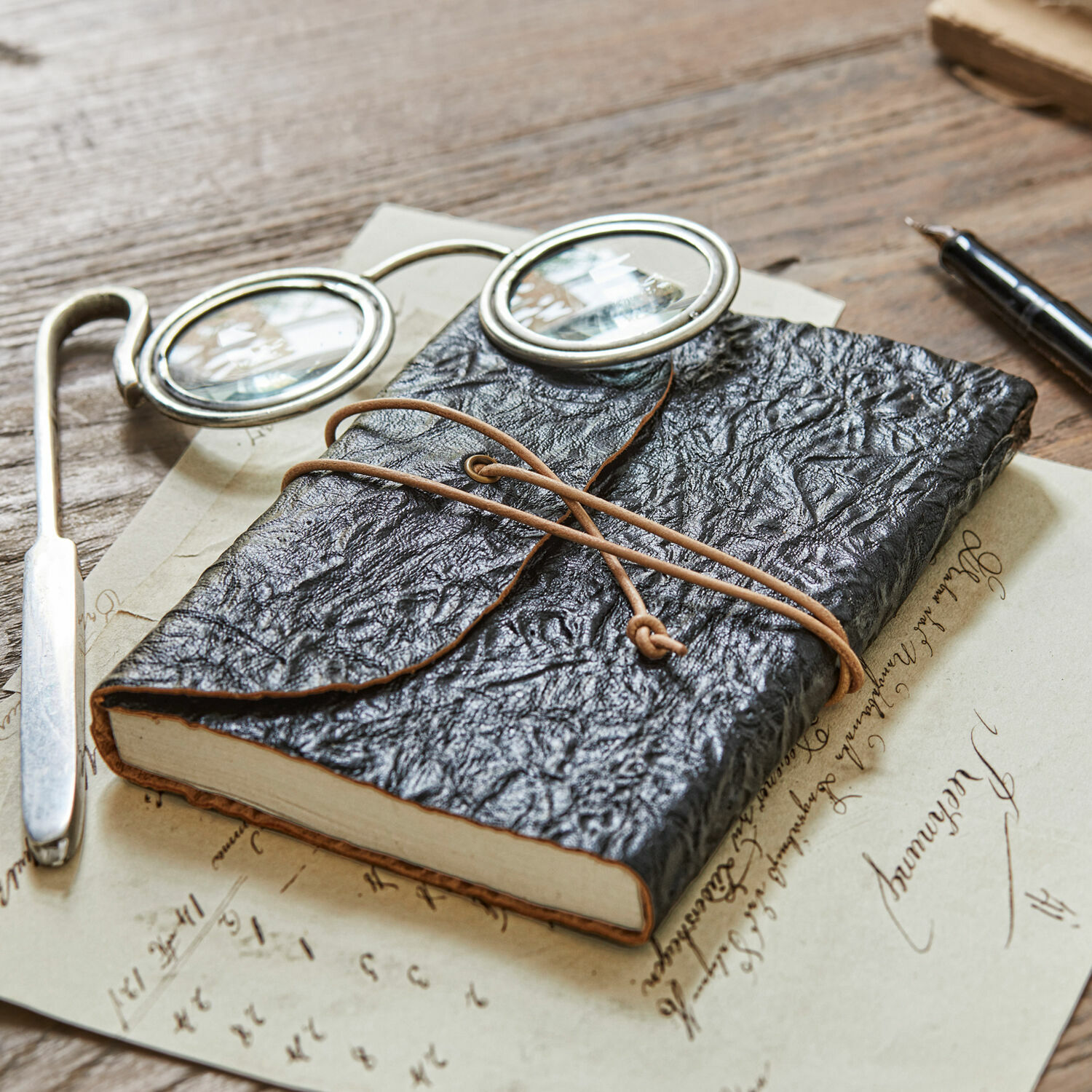 Notizbuch Cosmes Schwarzes Notizbuch aus strukturiertem Leder mit Lederriemen auf Holztisch mit Brille und Füller