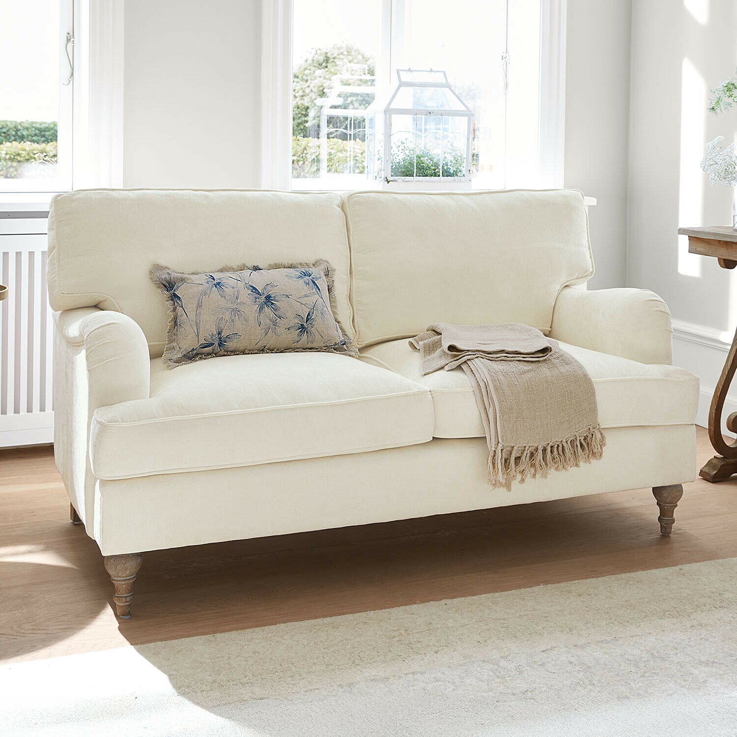 Sofa Elvika Beige bezogener Sofa mit floralem Kissen und beigem Plaid in hellem Wohnzimmer
