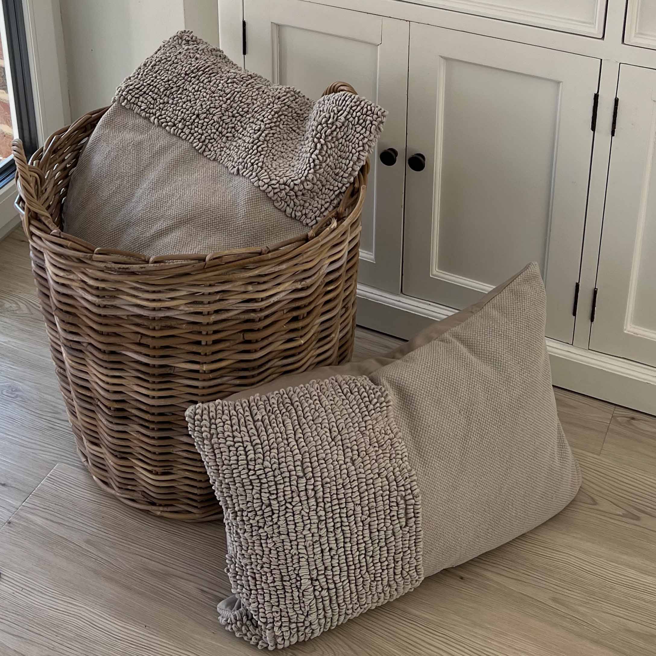 Beige Kissen aus weichem Material mit strukturierter Oberfläche neben einem Korb aus Weide auf Holzboden