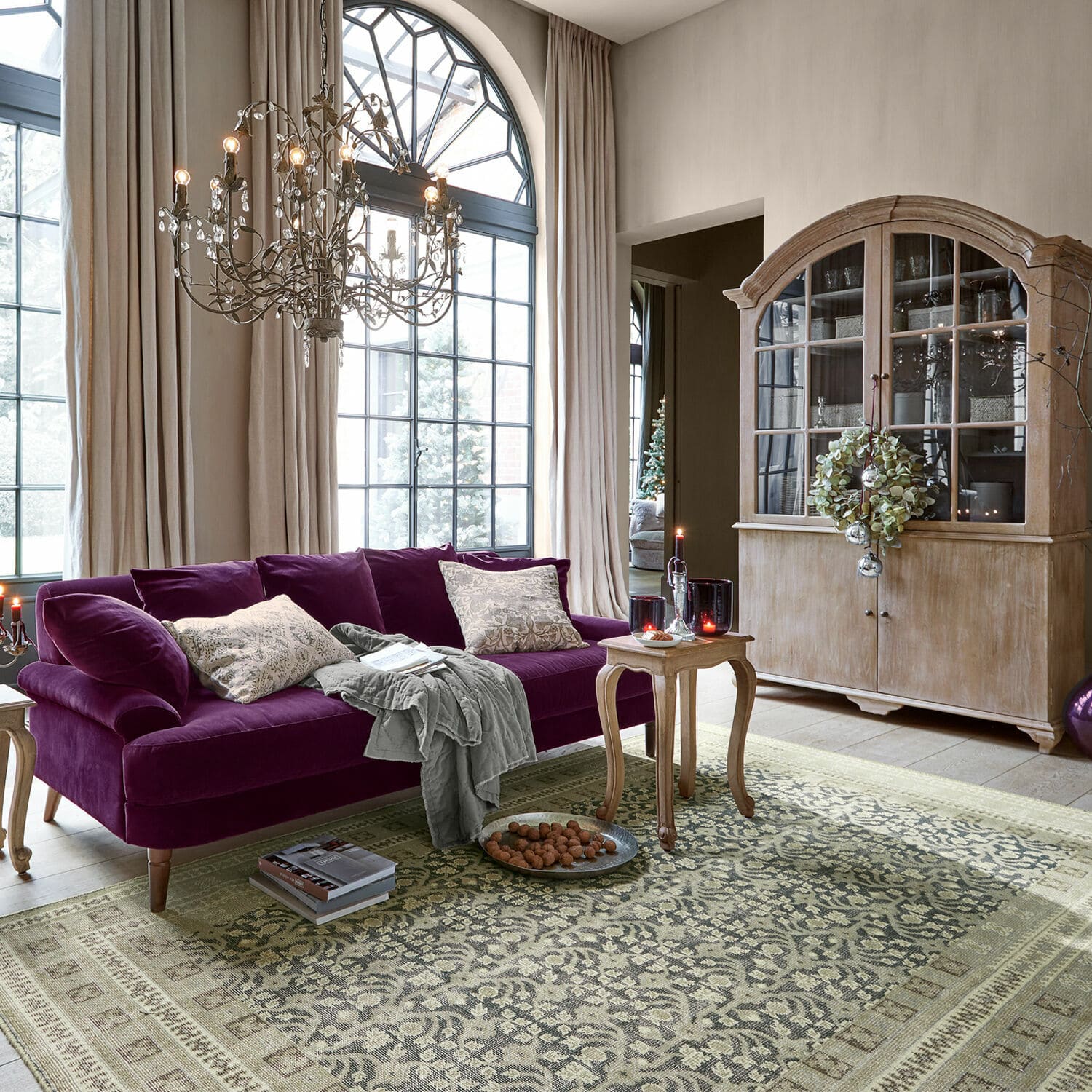 Wohnzimmer mit violettem Samtsofa und Holzvitrine vor großen Fenstern und gemustertem Teppich