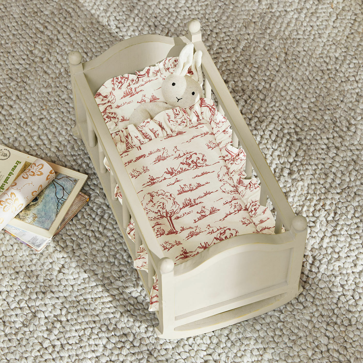 Puppenwiege Erilea Holz-Puppenbett in Creme mit rot gemusterter Bettwäsche und Plüschhase auf grauem Teppich