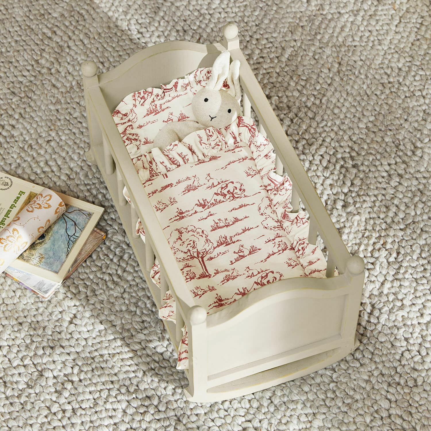 Puppenwiege Erilea Holz-Puppenbett in Creme mit rot gemusterter Bettwäsche und Plüschhase auf grauem Teppich