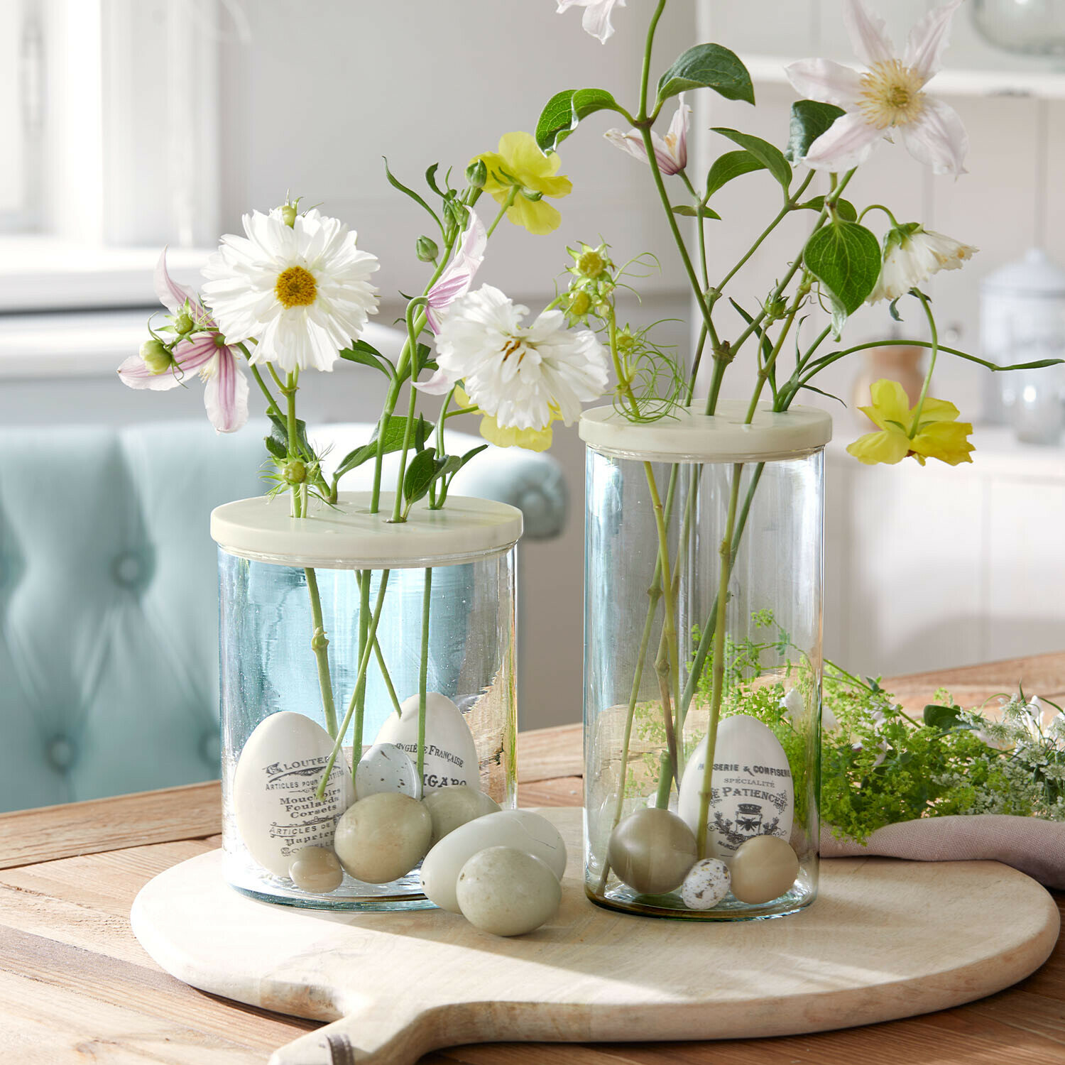 Glasvasen mit weißen und grünen Dekoeiern und Blumen auf einem Holzschneidebrett