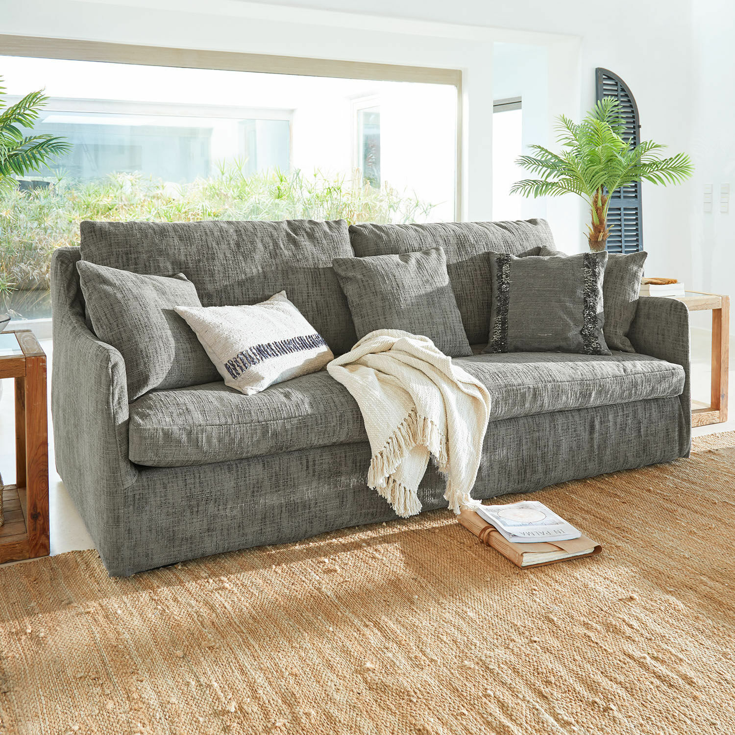 Sofa Shadowfield Graues Sofa aus Stoff mit Kissen und cremefarbener Decke auf Naturteppich in hellem Wohnzimmer