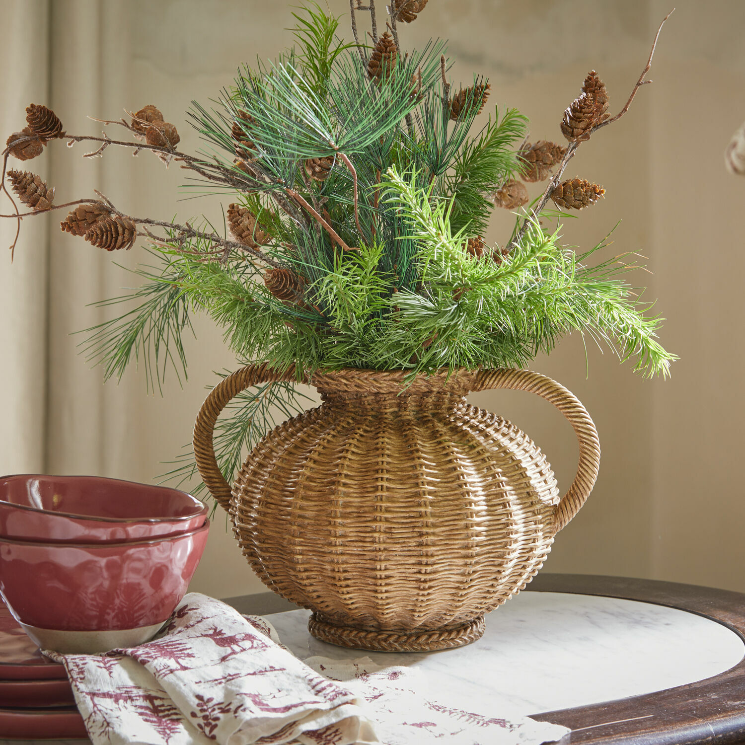 Vase Kaelvik Dekorativer Korb-Vase aus Rattan mit grünen Zweigen und Kiefernzapfen auf Tisch mit roter Keramik