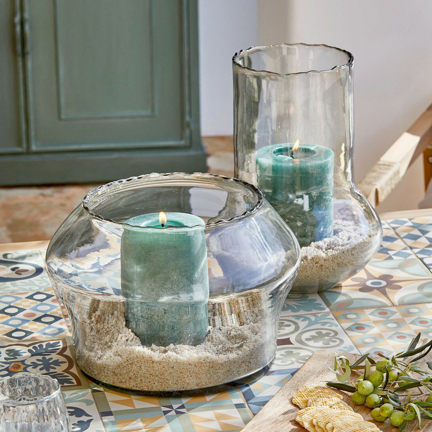 Windlicht 2er Set Lumiras Glaswindlichter mit grünen Kerzen und Sand auf einem bunten Tisch mit dekorativen Mustern