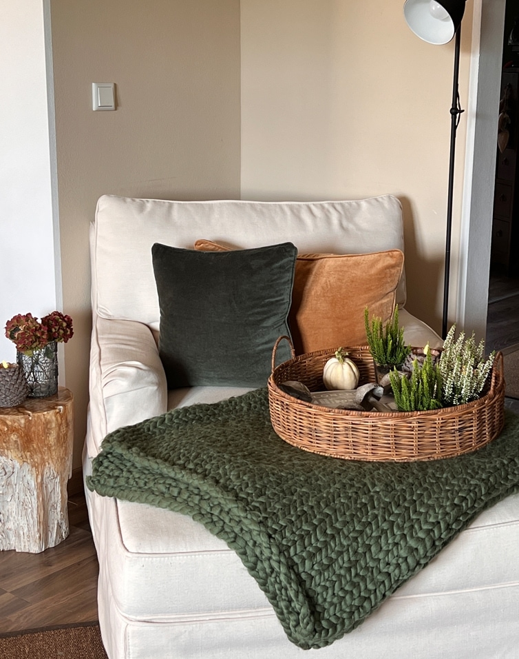 Beiger Sessel mit grünem und orangefarbenem Kissen, Korb mit Dekorationen auf grünem Strickplaid