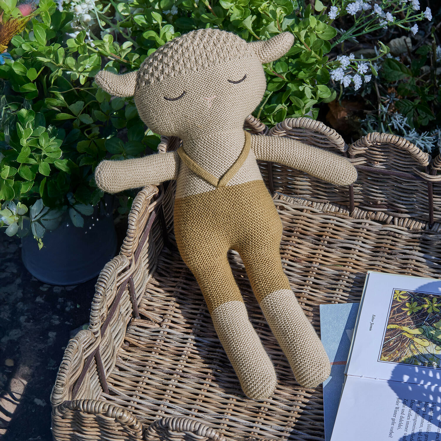Beige gestricktes Schaf Kuscheltier liegt im Weidenkorb neben Buch im Garten