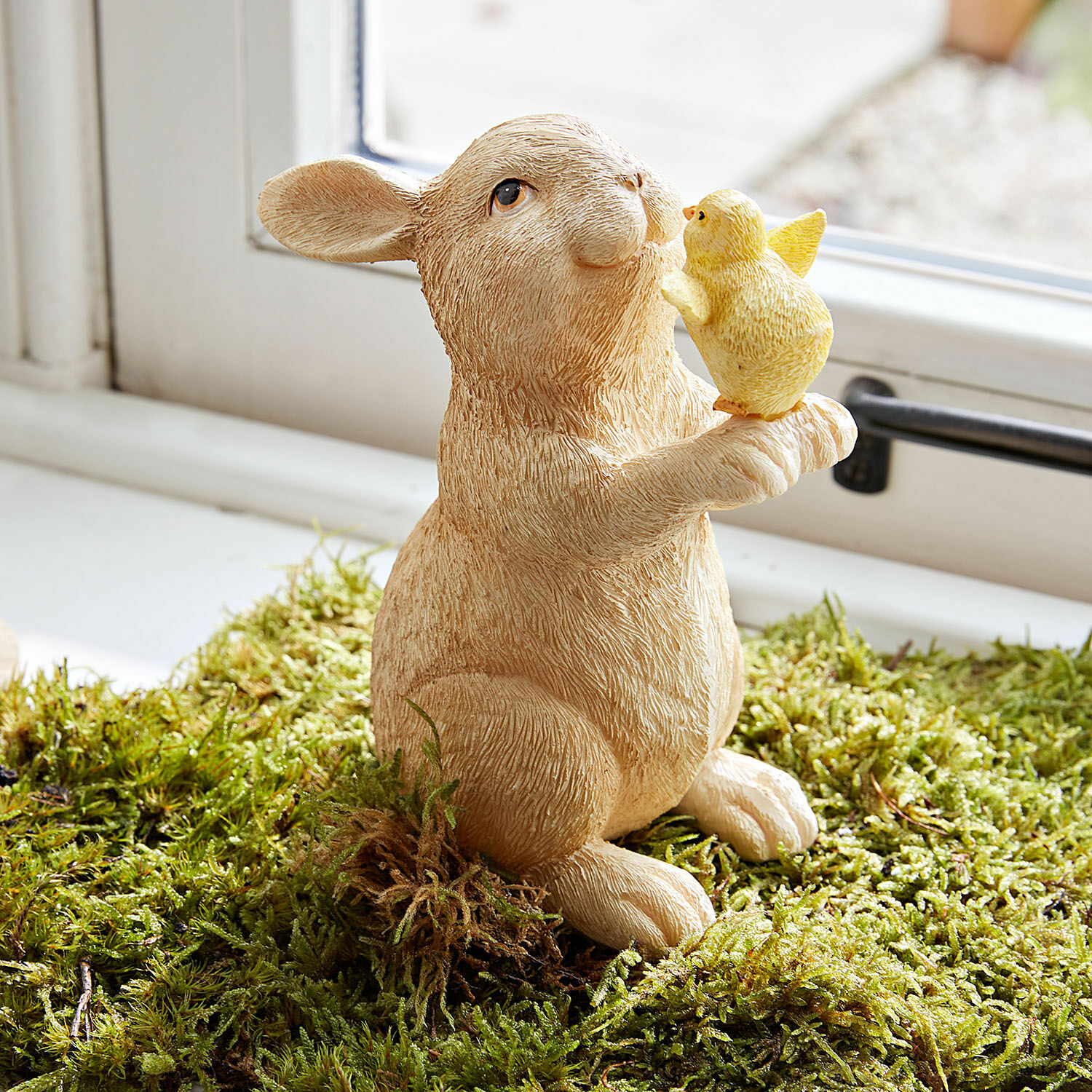 Deko-Hase Raniah Figur aus Harz mit beigem Hasen, der ein gelbes Küken auf grünem Moos hält, Frühlingsdekoration