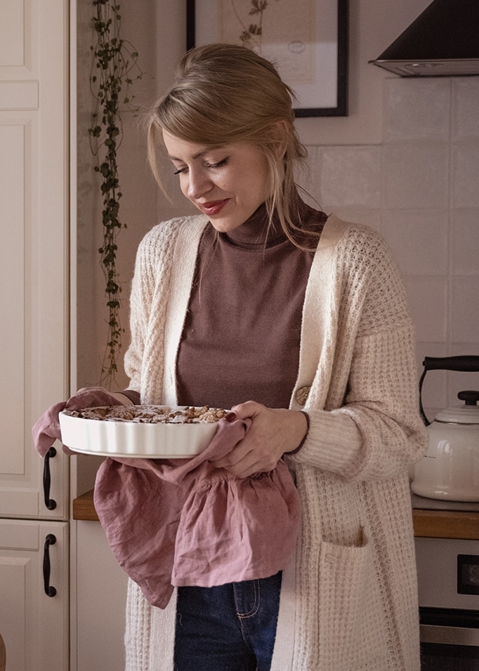 Frau im beigen Strickcardigan hält weiße Keramik-Backform mit rosa Tuch in der Küche