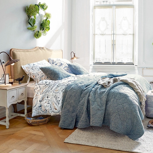 Gemütliches Schlafzimmer mit blauem Steppbett und gemustertem Bettwäsche-Set an beigem Kopfteil