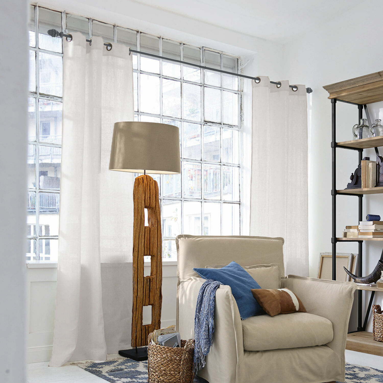 Gardine Salinas Stehlampe mit Holzfuß und beigem Lampenschirm in hellem Wohnzimmer mit Sessel und Regal