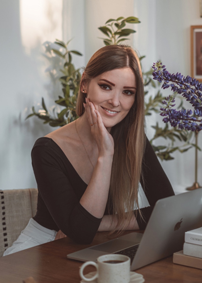 Frau sitzt an Holztisch mit Laptop, Kaffeetasse und Blumen im Hintergrund