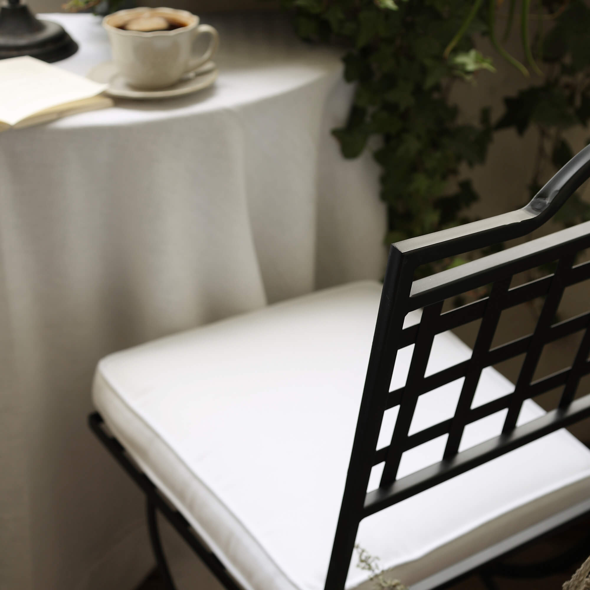 Schwarzer Metallstuhl mit weißem Sitzkissen vor Tisch mit Kaffeetasse und grünen Pflanzen auf Balkon