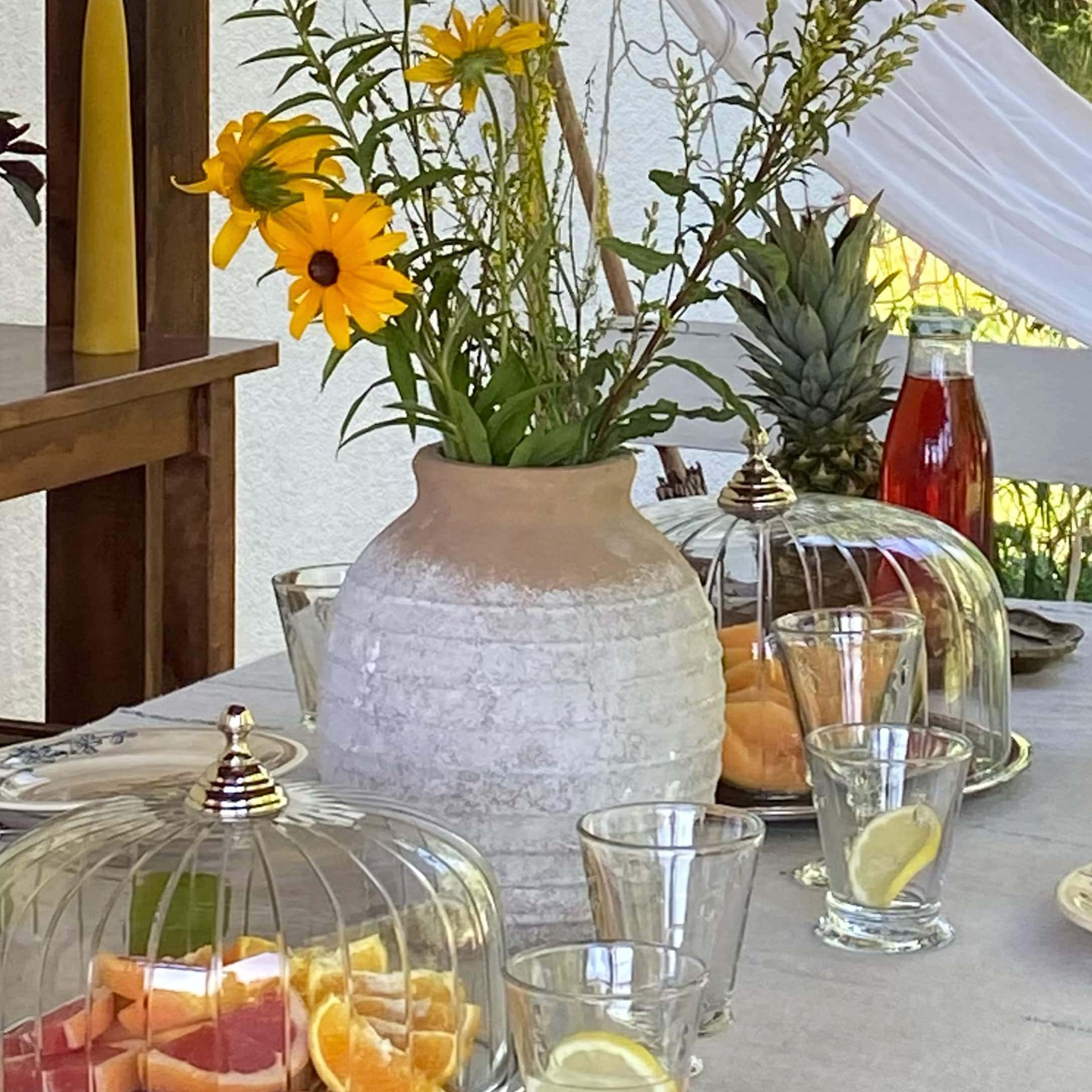 Keramikvase in Beige-Weiß mit gelben Blumen auf Tisch mit Früchten und Gläsern