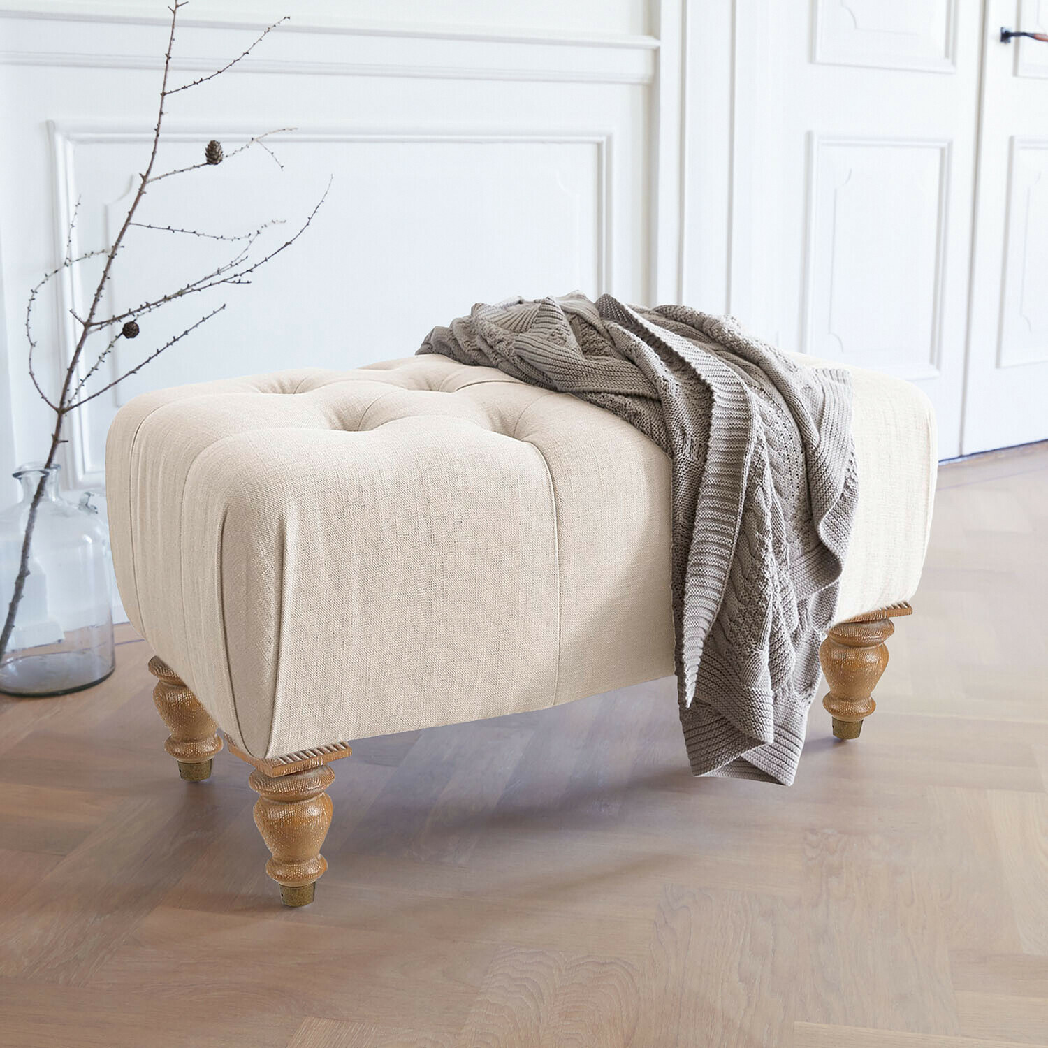 Hocker Durance Beiger gepolsterter Hocker mit Holzbeinen und grauer Strickdecke in hellem Raum