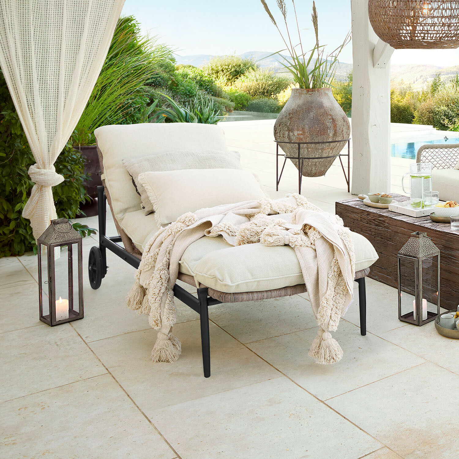 Gartenliege mit cremefarbenen Kissen und weicher Decke auf Terrasse mit Poolblick und grüner Umgebung
