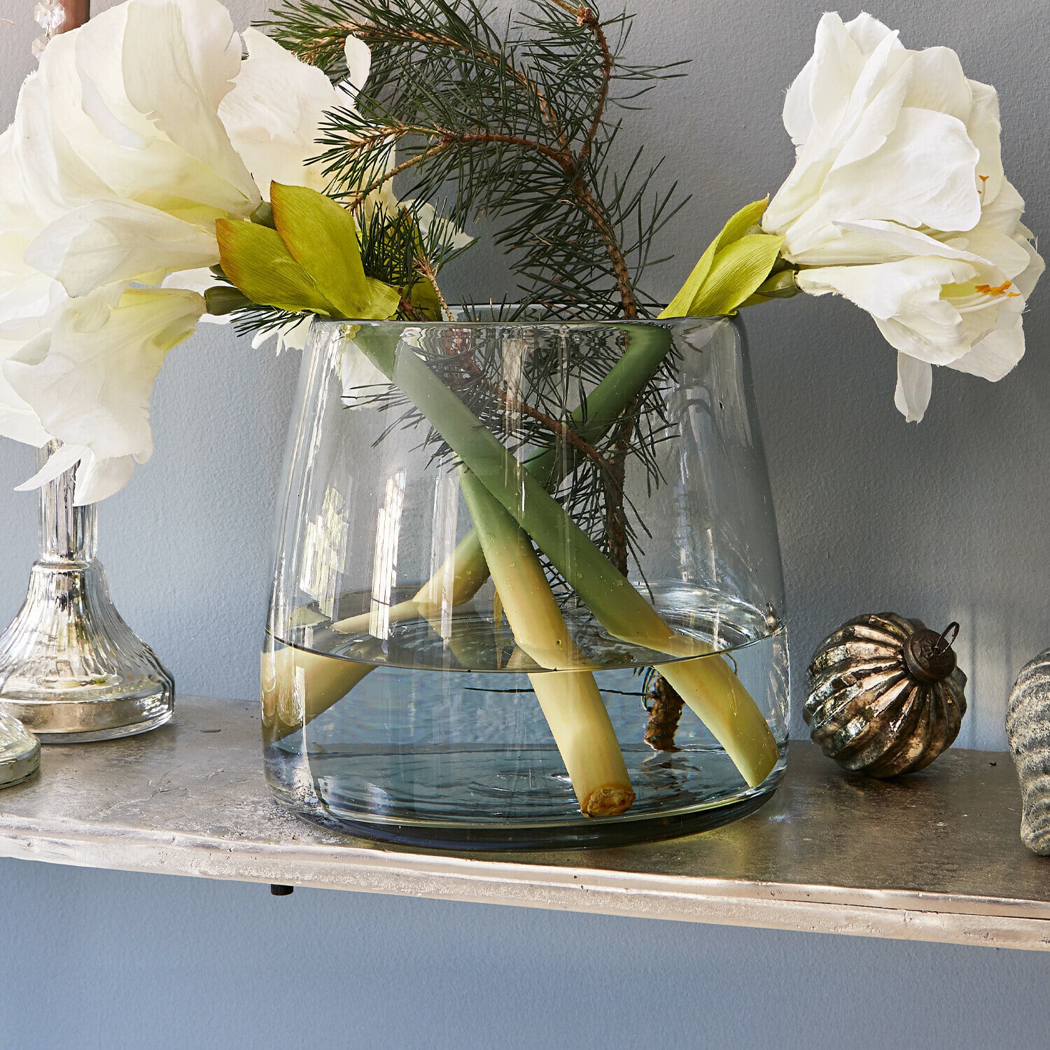 Vase Inga Transparente Glasvase mit weißen Blumen und grünen Zweigen auf einem dekorativen Regal