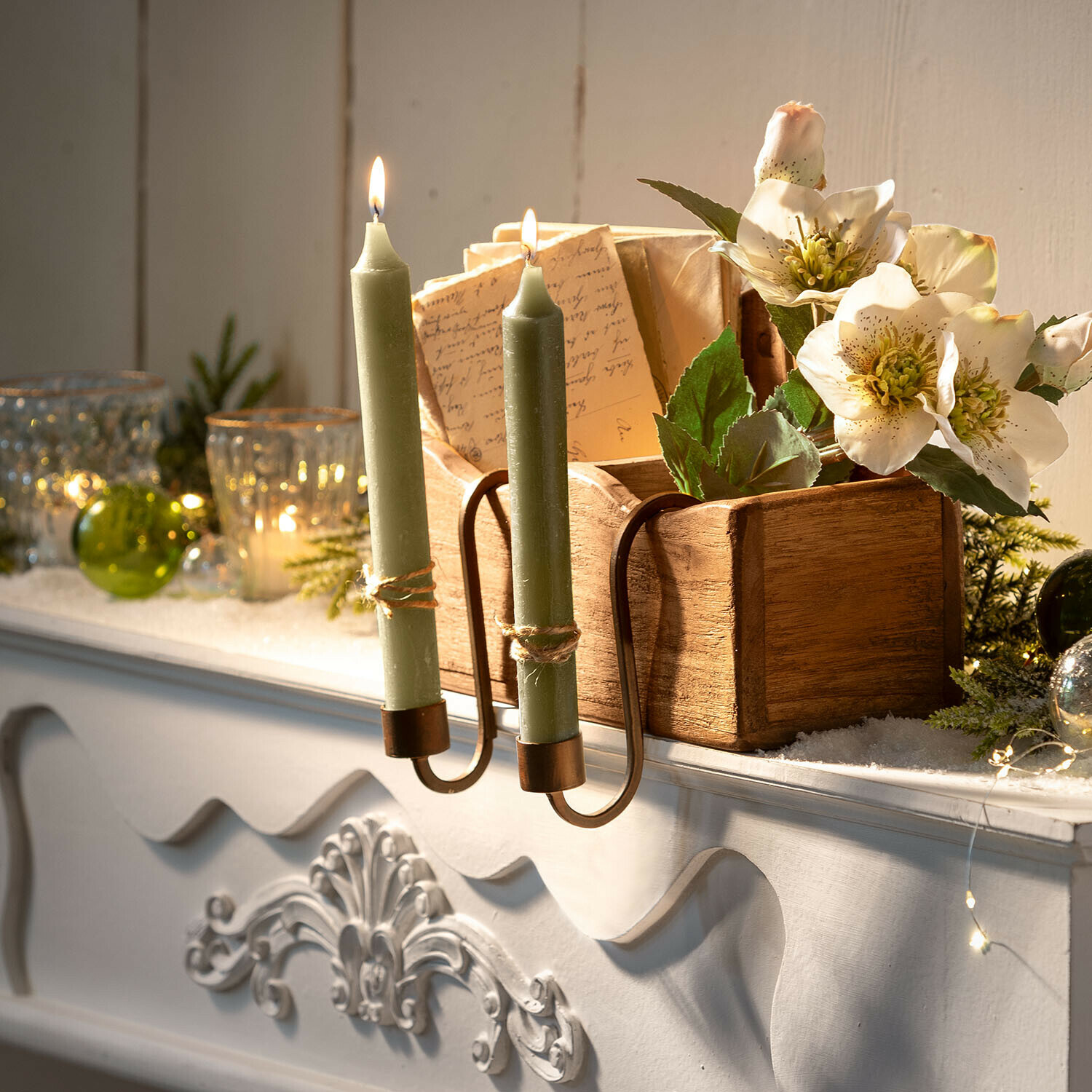 Doppelter Messing-Kerzenhalter mit grünen Kerzen auf weißem Kaminsims mit Blumen und Holzkiste