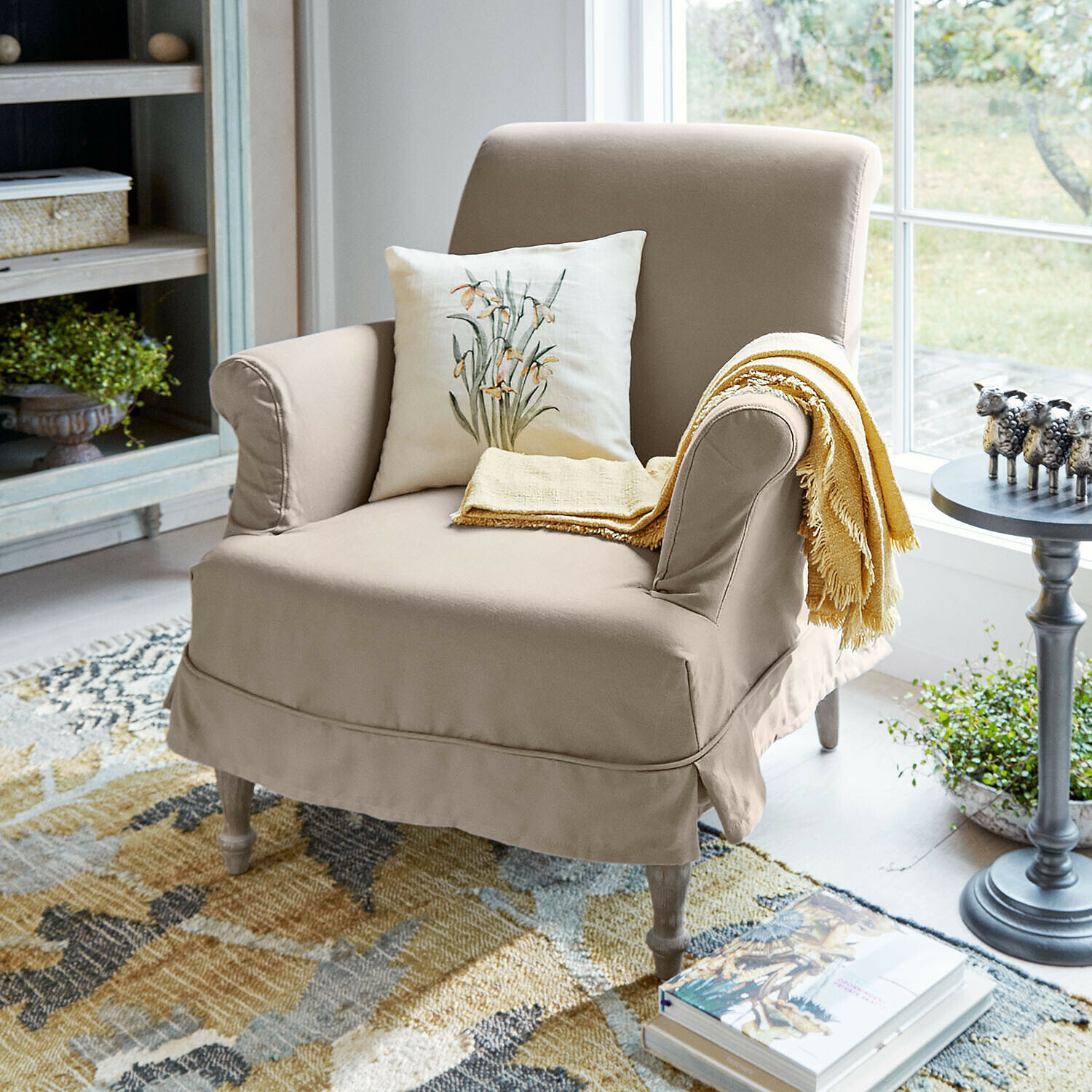 Sessel Everleighs Beiger Polsterstuhl aus Stoff mit floralem Kissen und gelbem Plaid in hellem Wohnzimmer mit Teppich