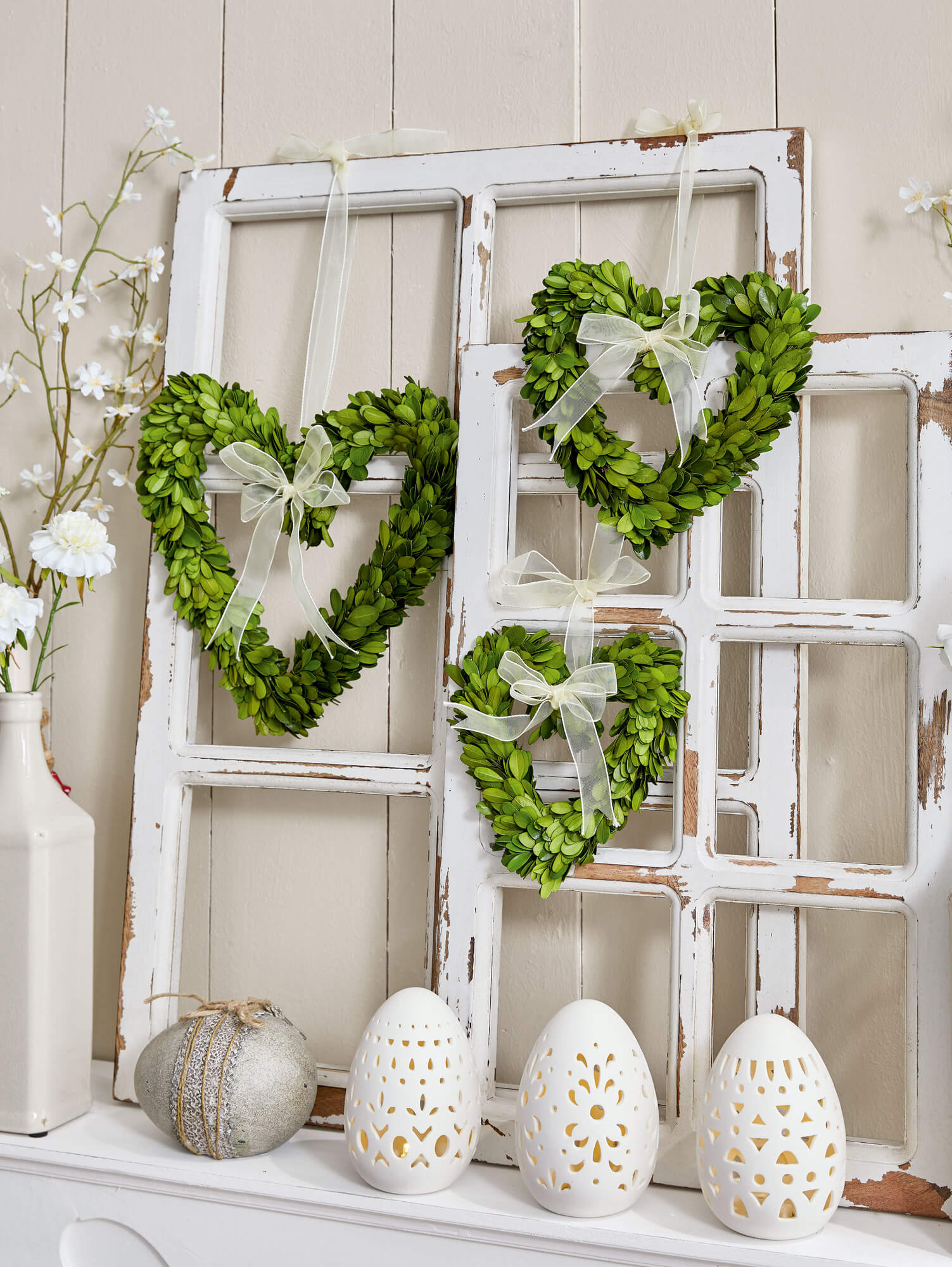 Grüne Herzkränze aus Blättern mit cremefarbenen Schleifen an weißen, shabby-chic Fensterrahmen mit dekorativen Eiern