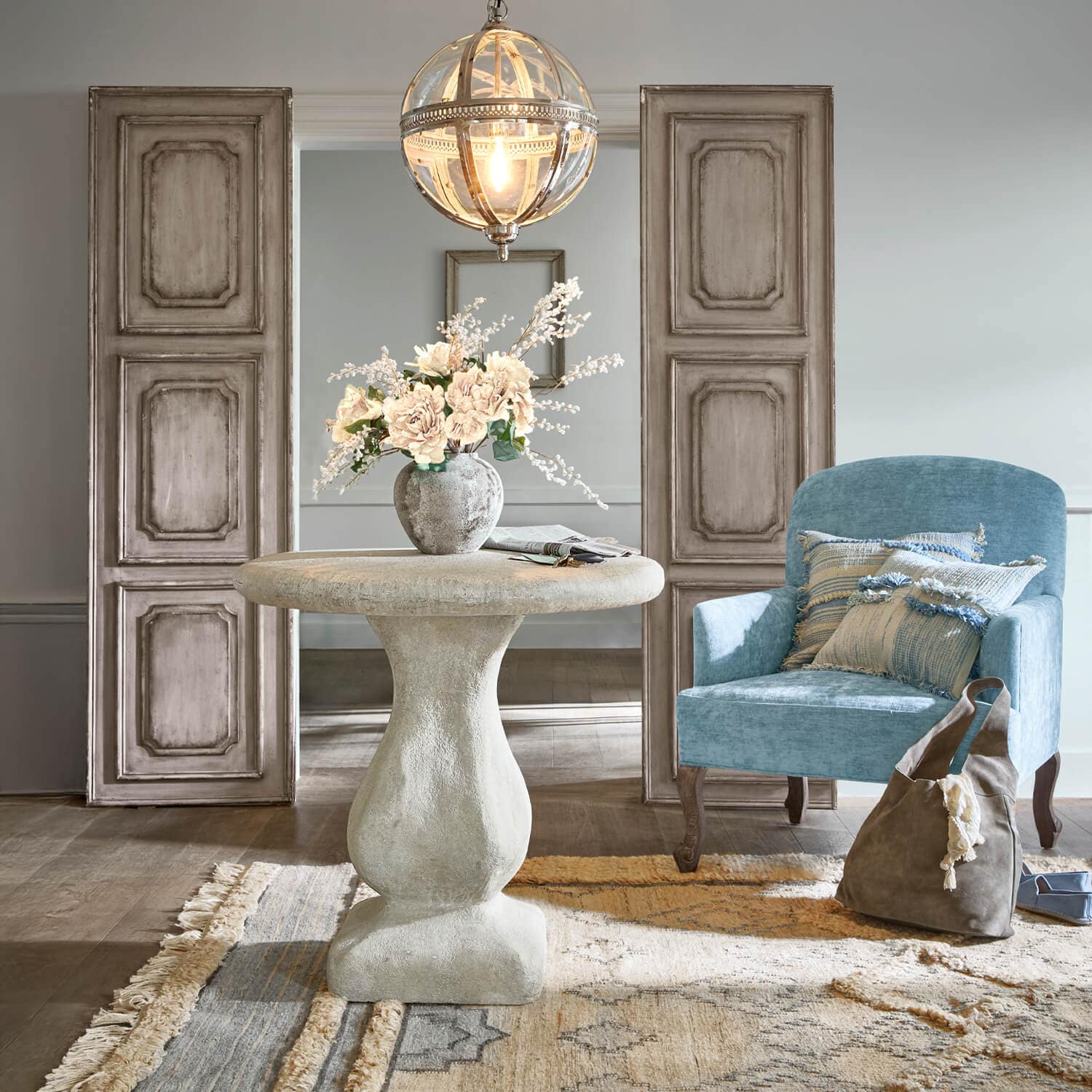 Runder grauer Betontisch mit Blumenvase, blauer Sessel und braune Tasche in hellem Raum