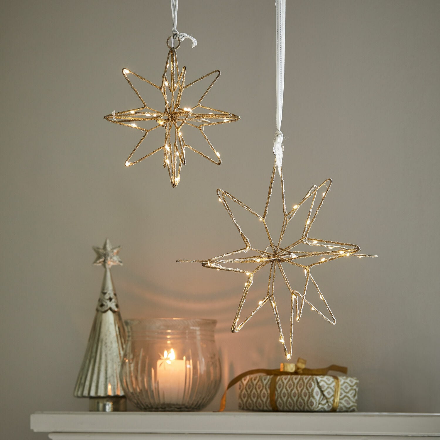 LED-Stern 2er Set Pérole Goldene LED-Stern-Dekorationen hängen über Kerze und Geschenk auf weißem Regal