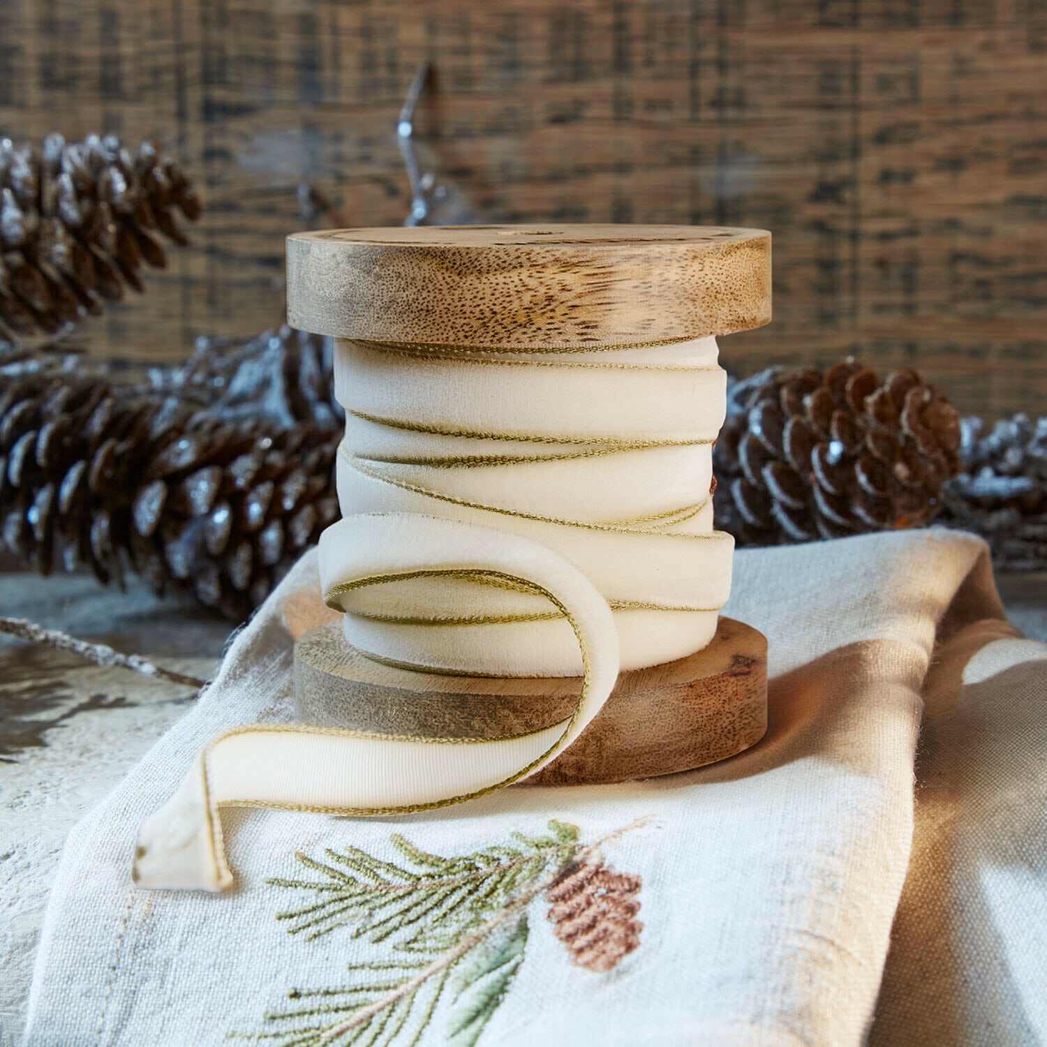 Samtband Sorima Natürliche Holzspule mit cremefarbenem Band und grünem Rand auf Leinenstoff mit Kiefernzapfen-Stickerei