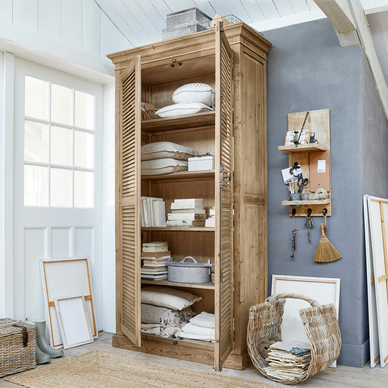 Schrank Farnham Holzschrank mit Lamellentüren in Naturholzfarbe, gefüllt mit Kissen und Büchern in hellem Raum