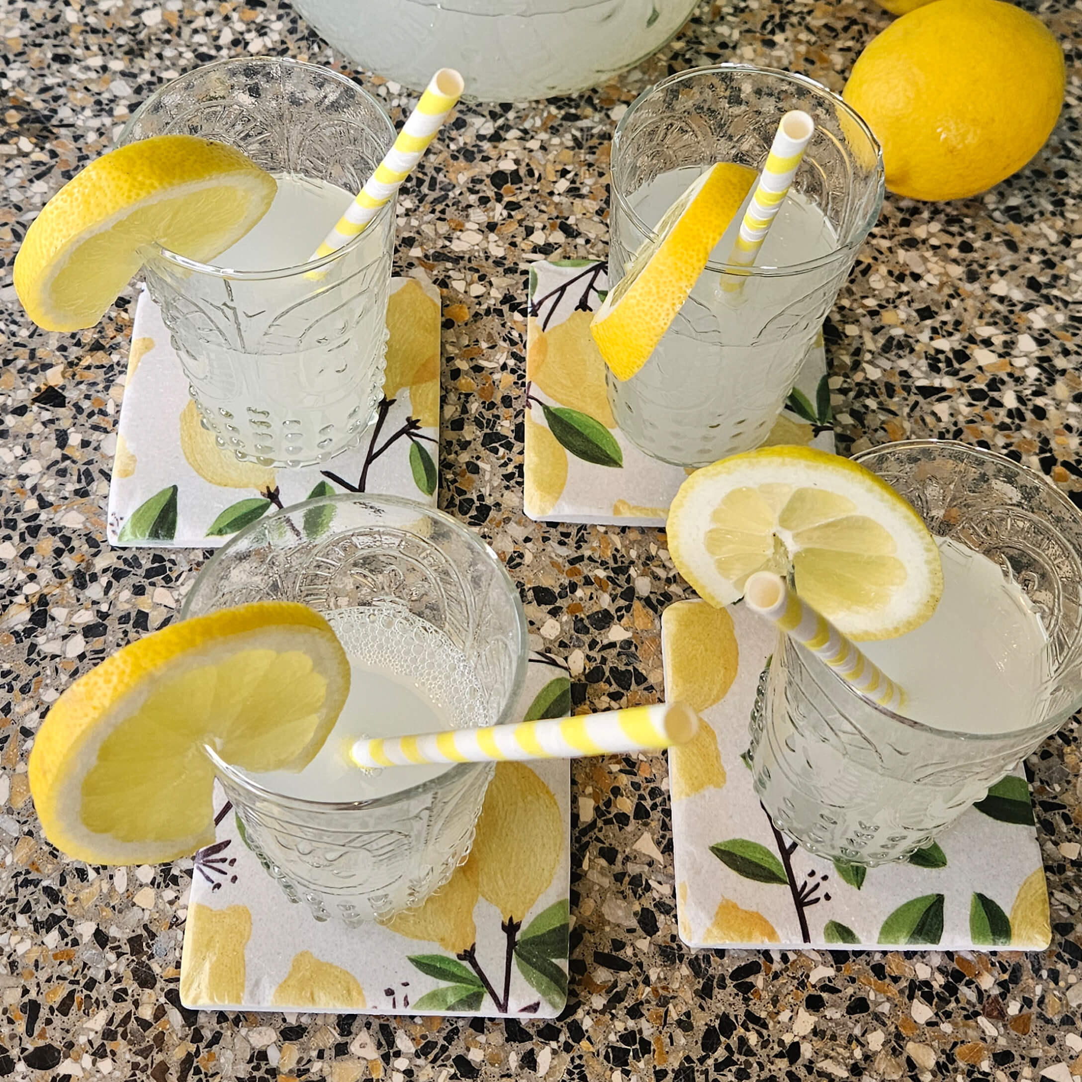 Gläser mit Limonade und Zitronenscheiben auf Keramikuntersetzern mit Zitronenmuster auf Steinplatte