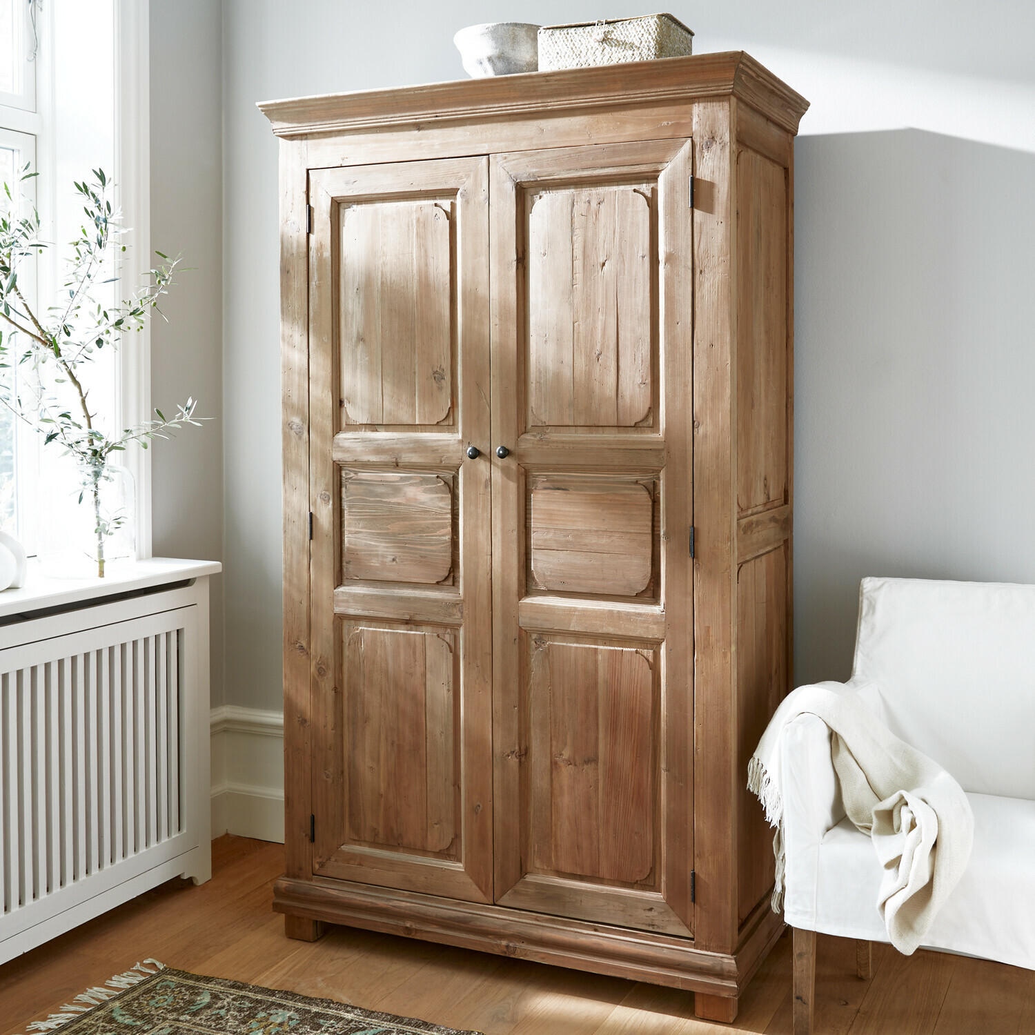 Kleiderschrank Gavrielle Naturfarbener Holzschrank in hellem Raum mit weißem Sessel und gemustertem Teppich