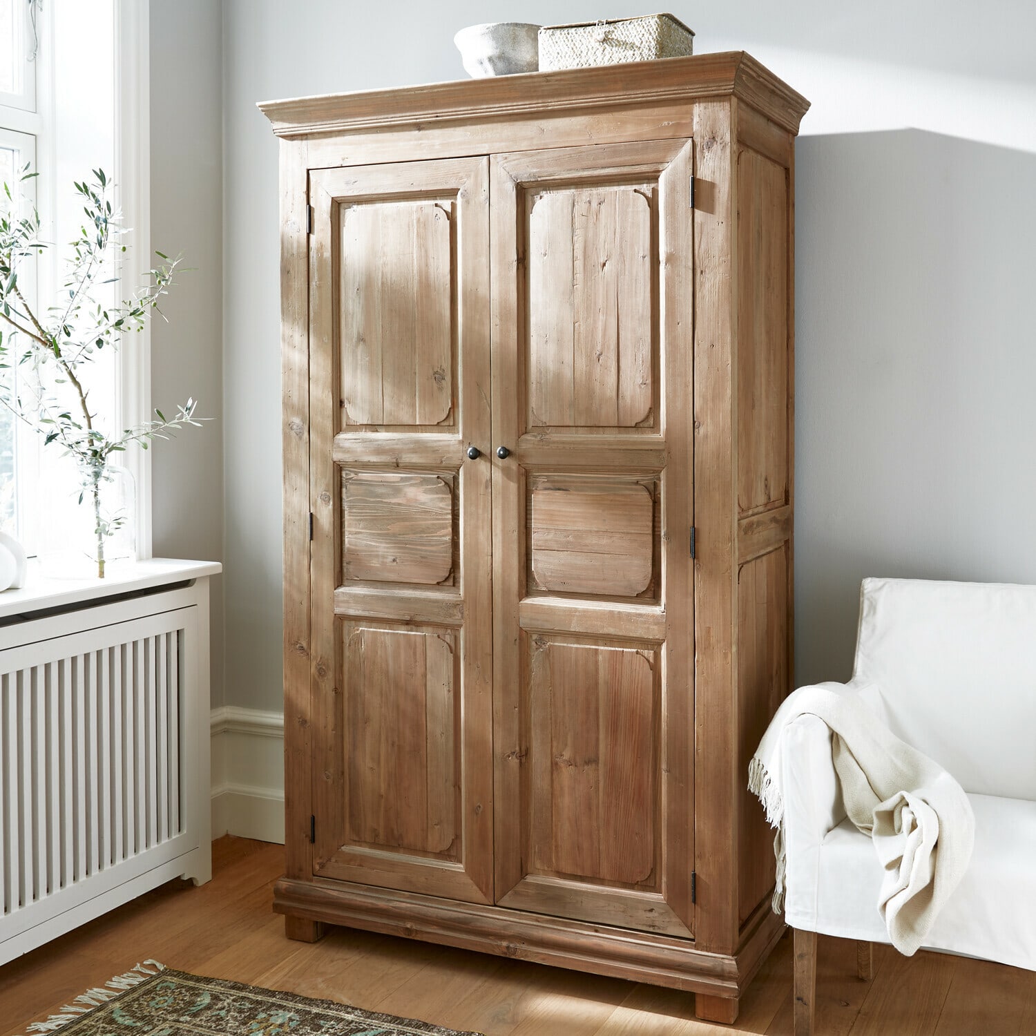 Kleiderschrank Gavrielle Naturfarbener Holzschrank in hellem Raum mit weißem Sessel und gemustertem Teppich