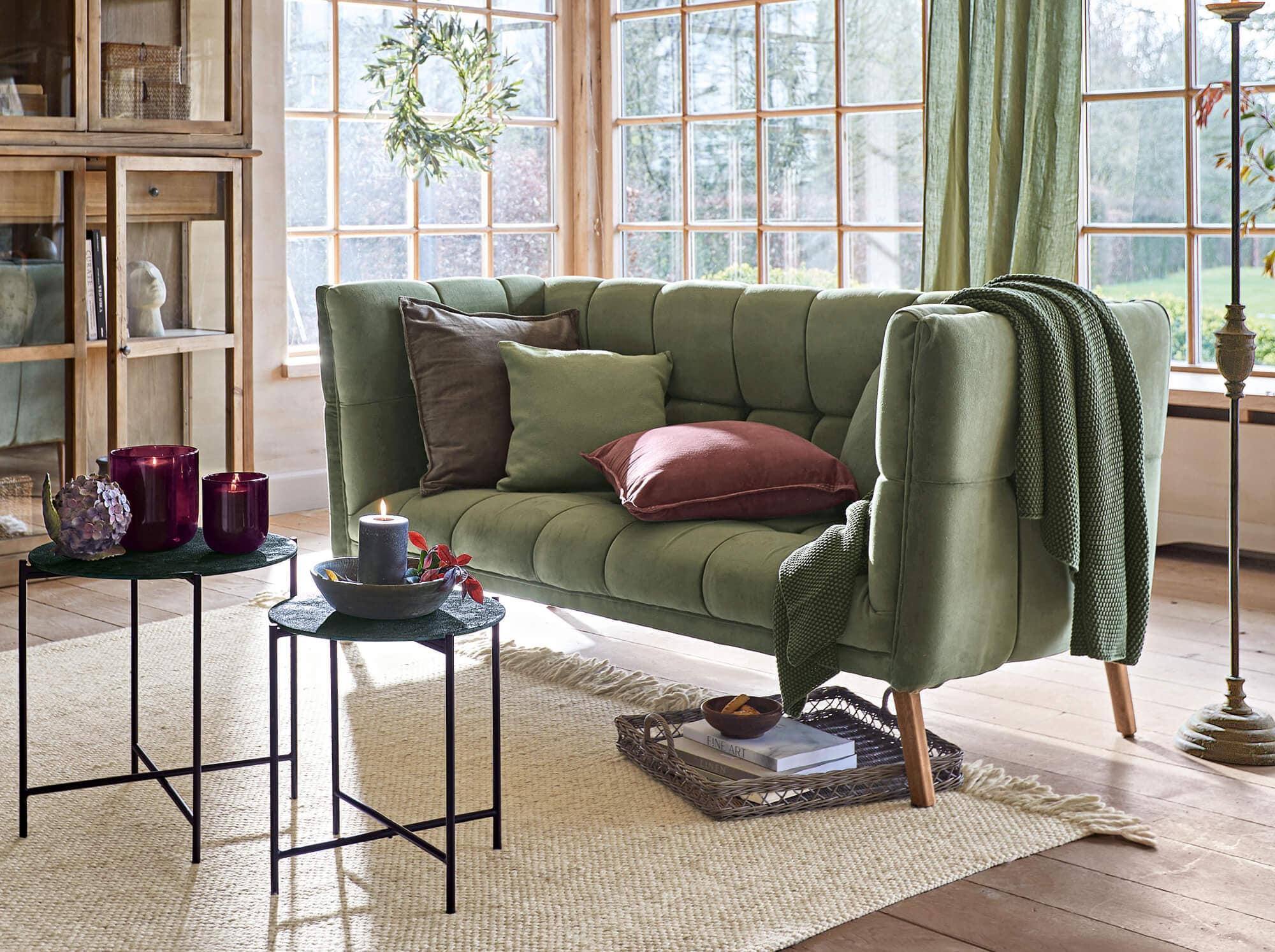 Grünes Samt-Vintage-Sofa mit Kissen und Decke in hellem Wohnzimmer mit großen Fenstern
