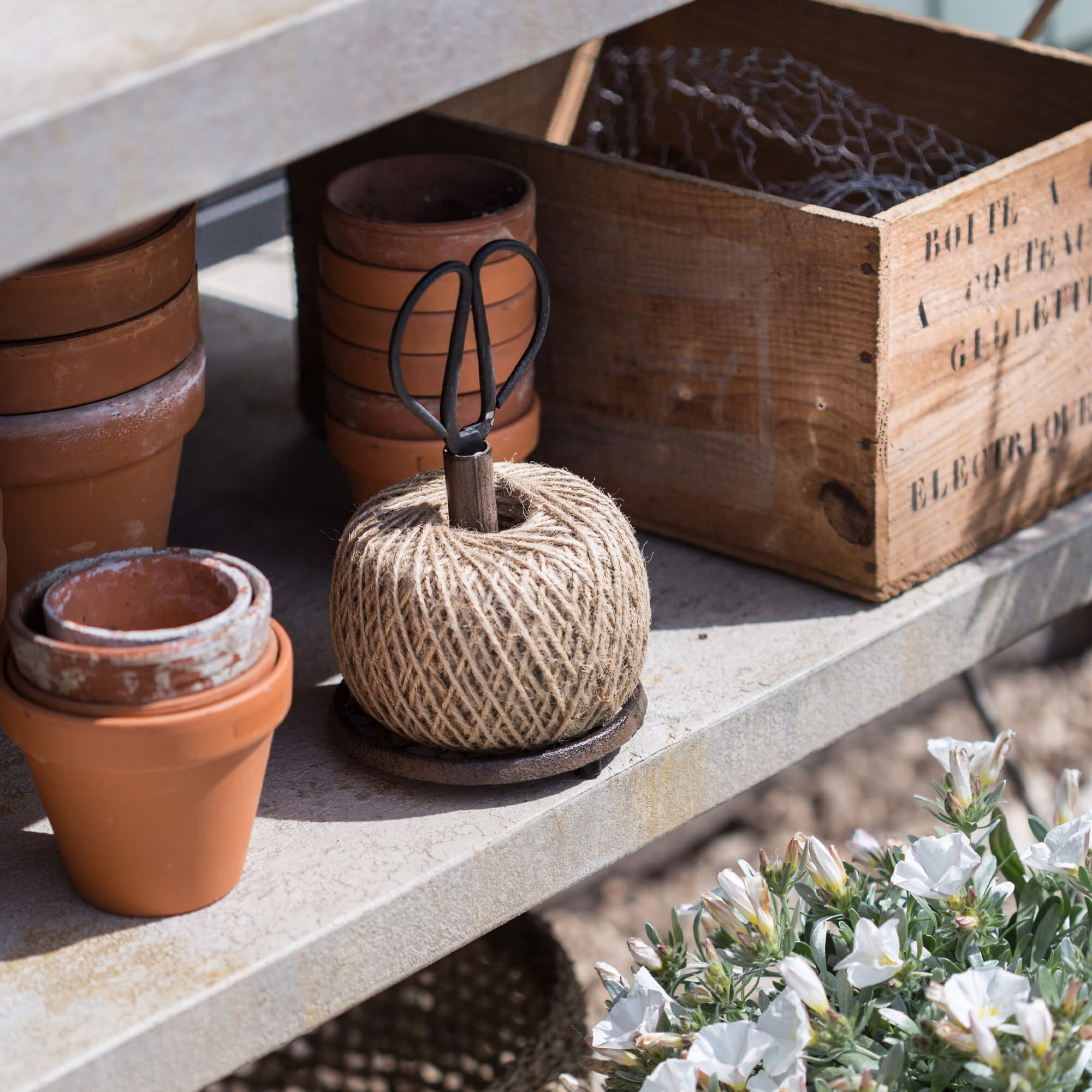 Natürliche Gartenschnur mit Metallschere auf Betonschale neben Terrakottatöpfen und Holzkiste