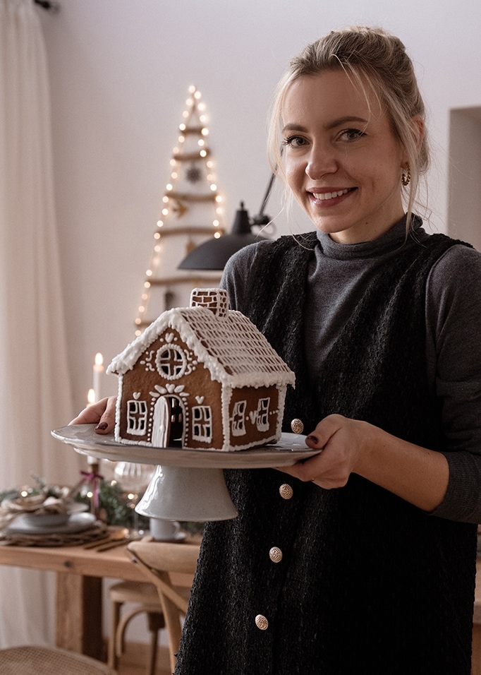 Frau hält einen Teller mit einem Lebkuchenhaus in gemütlich weihnachtlich dekoriertem Raum