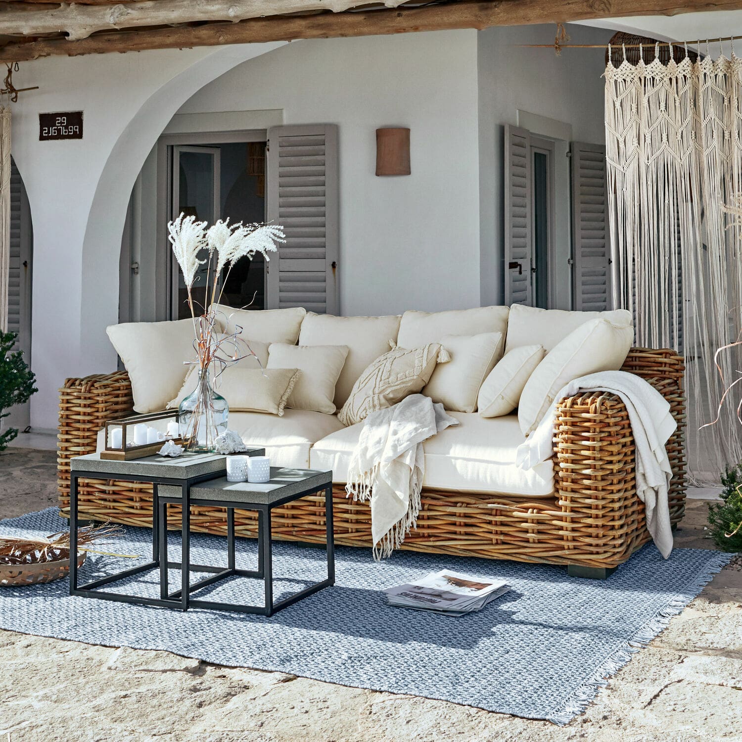 Sofa Rimini Rattansofa mit cremefarbenen Kissen und Decke auf blauem Teppich auf sonniger Terrasse