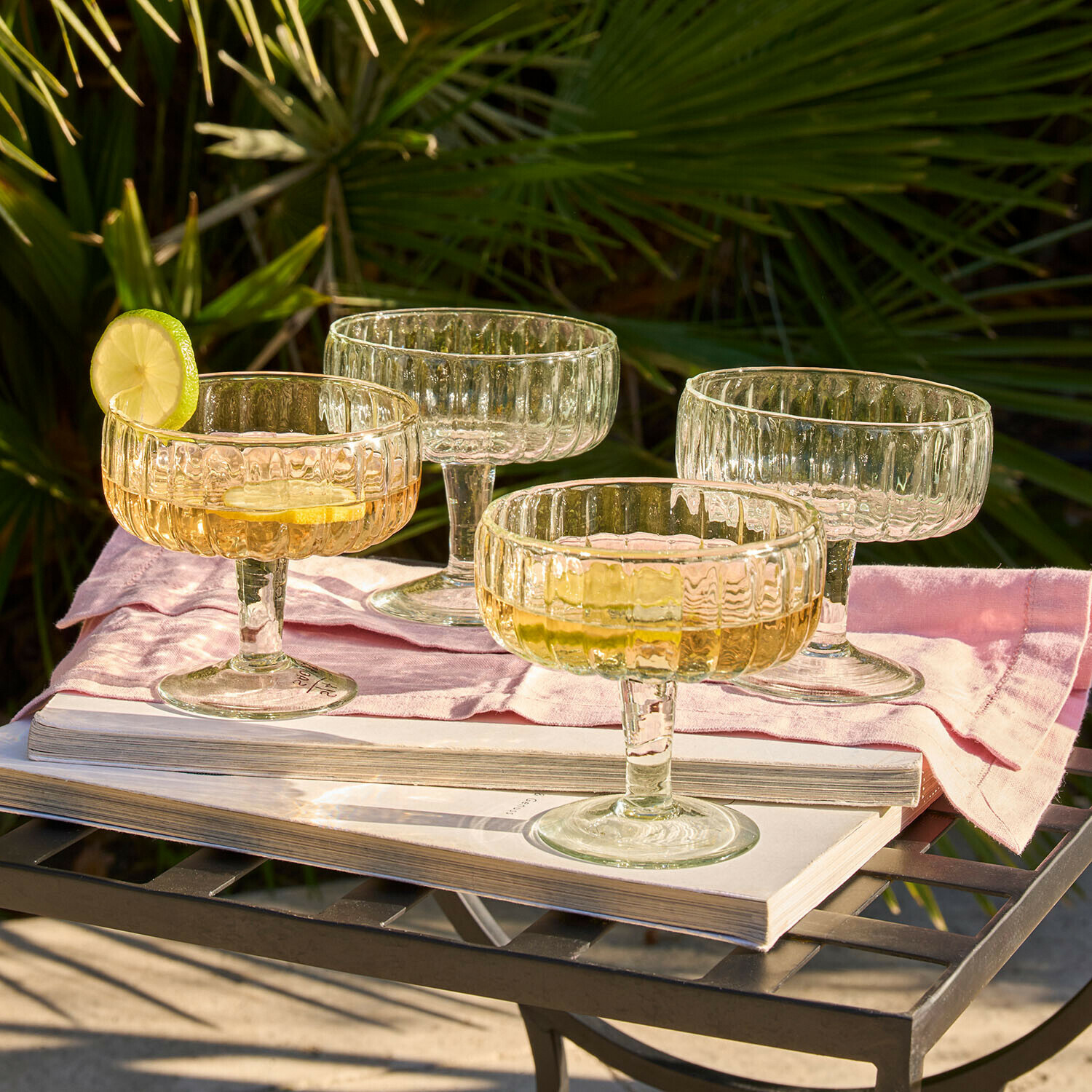 Glas 4er Set Solerón Set aus vier transparenten Cocktailgläsern aus geriffeltem Glas auf Metalltisch mit Buch und rosa Serviette
