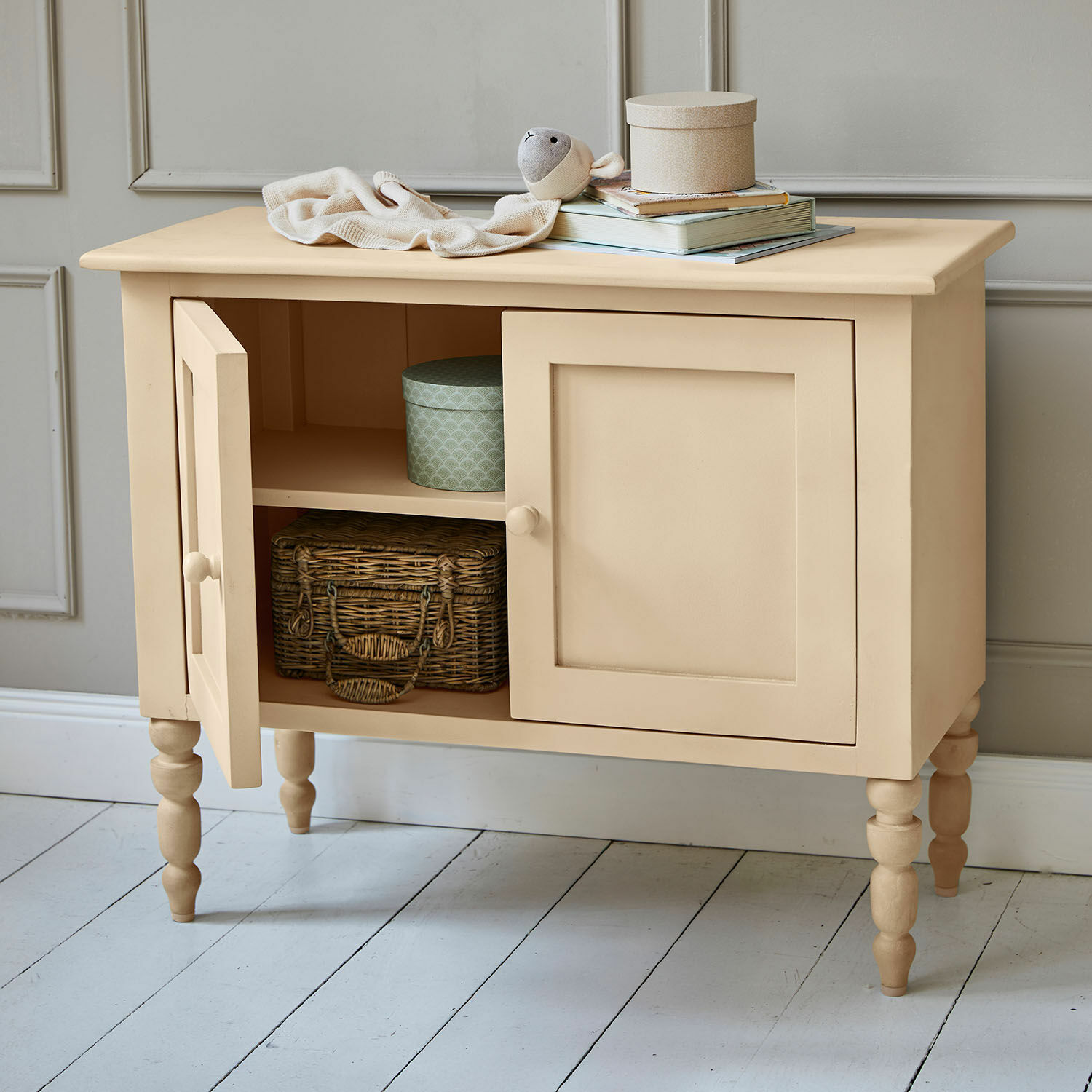 Beige lackierte Holzkommode mit zwei Türen und gedrechselten Beinen, mit Korb und Dosen im Regal