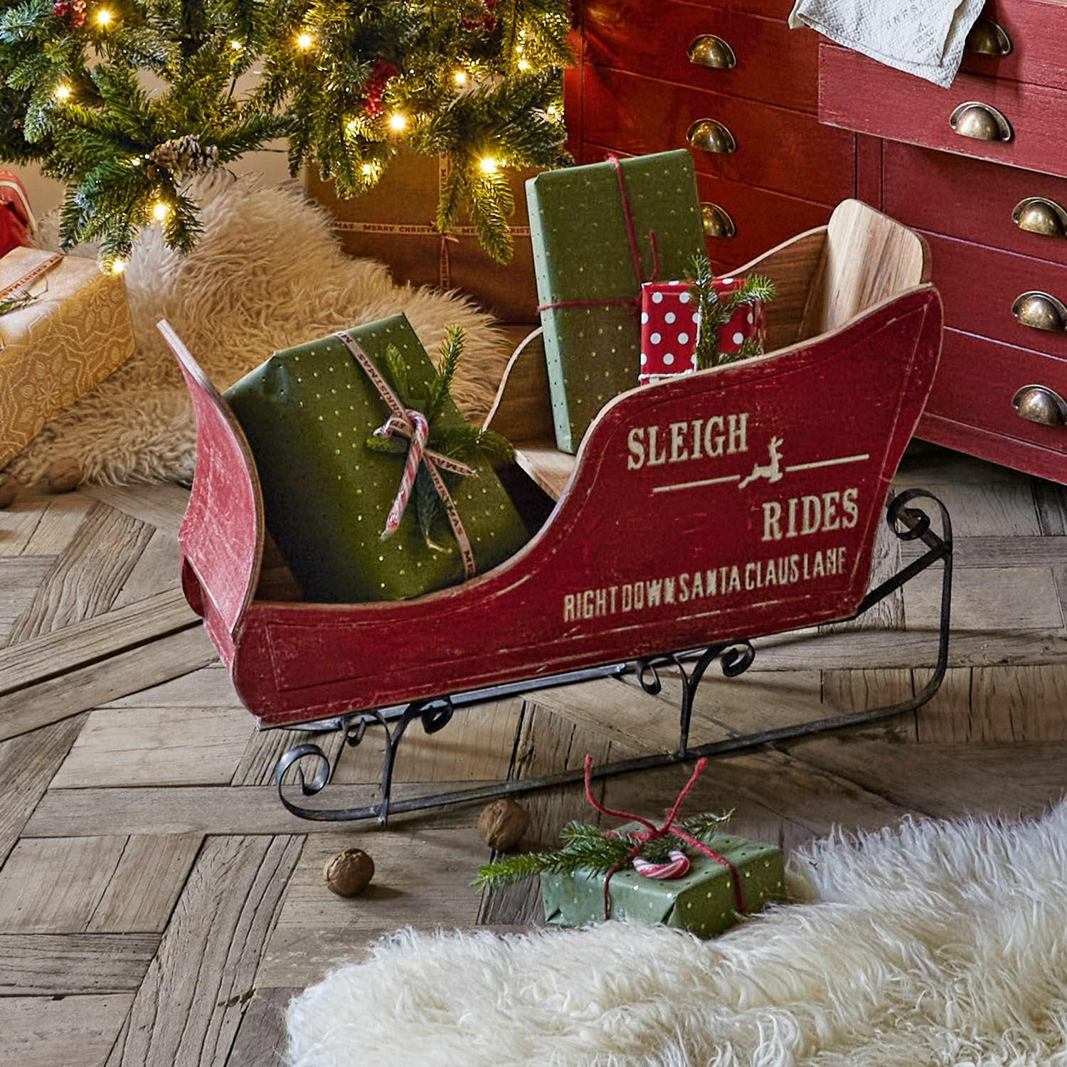 Deko-Schlitten Maélys Roter Holzschlitten mit Metallkufen und grünen Geschenken auf Holzboden neben geschmücktem Weihnachtsbaum
